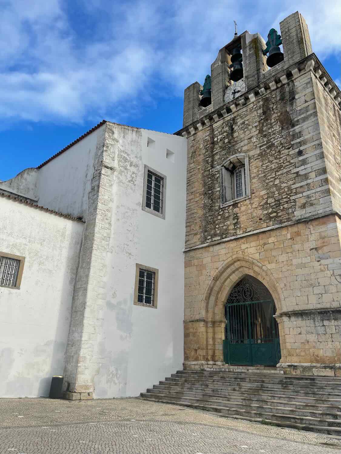 The imposing architecture of a church in Faro, featuring a tall bell tower with green rooftop accents, a Gothic arched entrance, and a contrast of white walls against the blue sky.