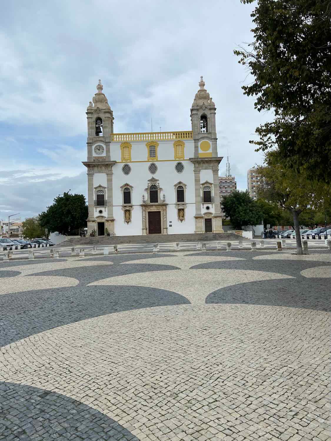 Historic Faro church with striking white facade and twin bell towers, set against a patterned cobblestone square on a cloudy day in Faro, as seen during a Lisbon day trip."

