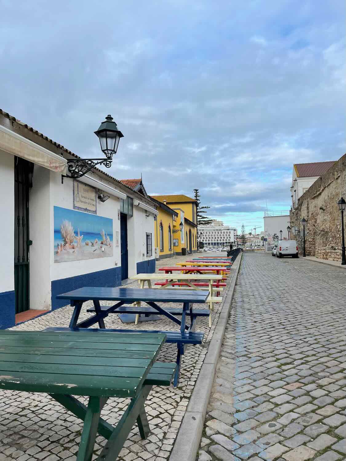 Cobblestone street lined with colorful picnic tables and white buildings with blue accents under a lamp post, in downtown Tavira.