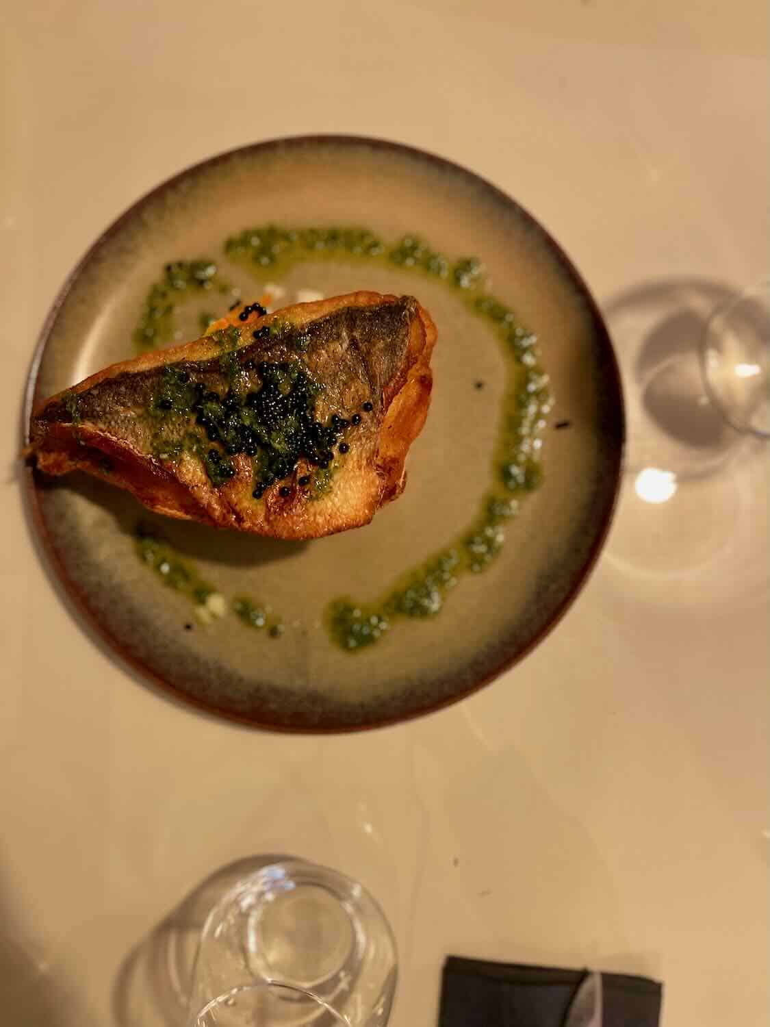 A plate of grilled fish topped with a green herb sauce, presented on a ceramic plate with decorative green sauce at a restaurant in Tavira.