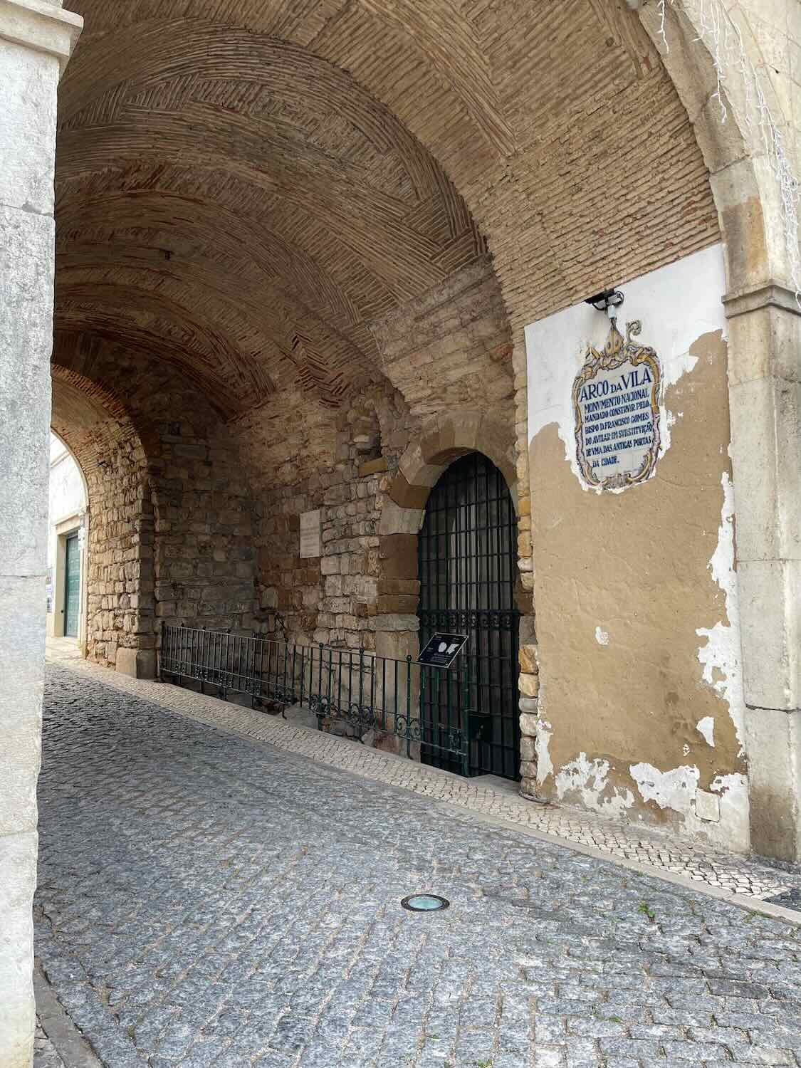 Arched stone gateway known as 'Arco da Vila' in Faro, with intricate brickwork ceiling and a historical plaque on the peeling plaster wall; cobblestone pavement adds to the historical ambiance.