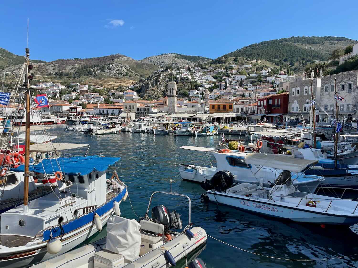 Vibrant Hydra harbor scene bustling with boats and yachts, framed by the picturesque hillside architecture of the island, under the clear blue Greek sky.