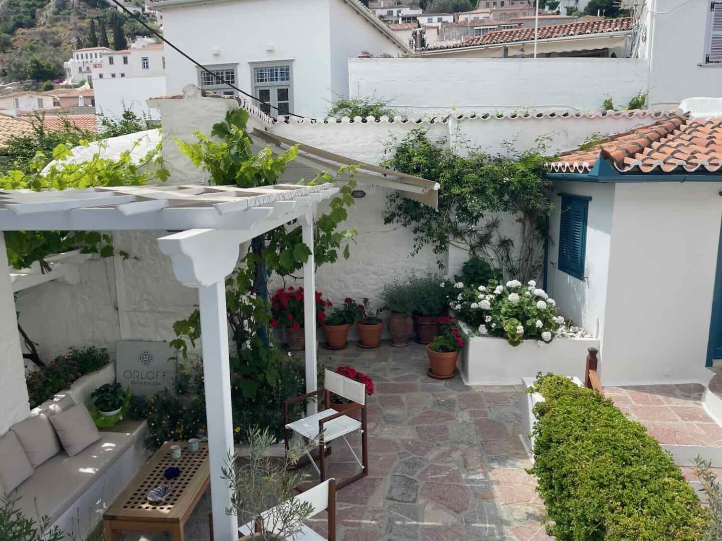 Charming patio of Orloff in Hydra, lined with flowering pots and a pergola, providing a peaceful spot for guests to unwind amidst the island's beauty.