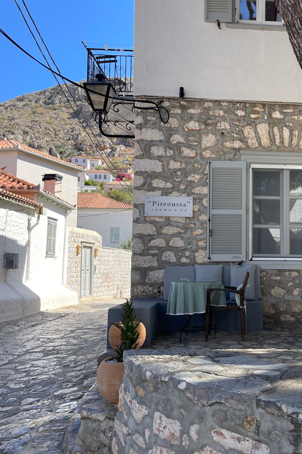 Quaint cobblestone alley in Hydra leading to 'Piteoussa rooms & co,' a charming guesthouse with inviting outdoor seating, nestled among traditional Greek architecture.