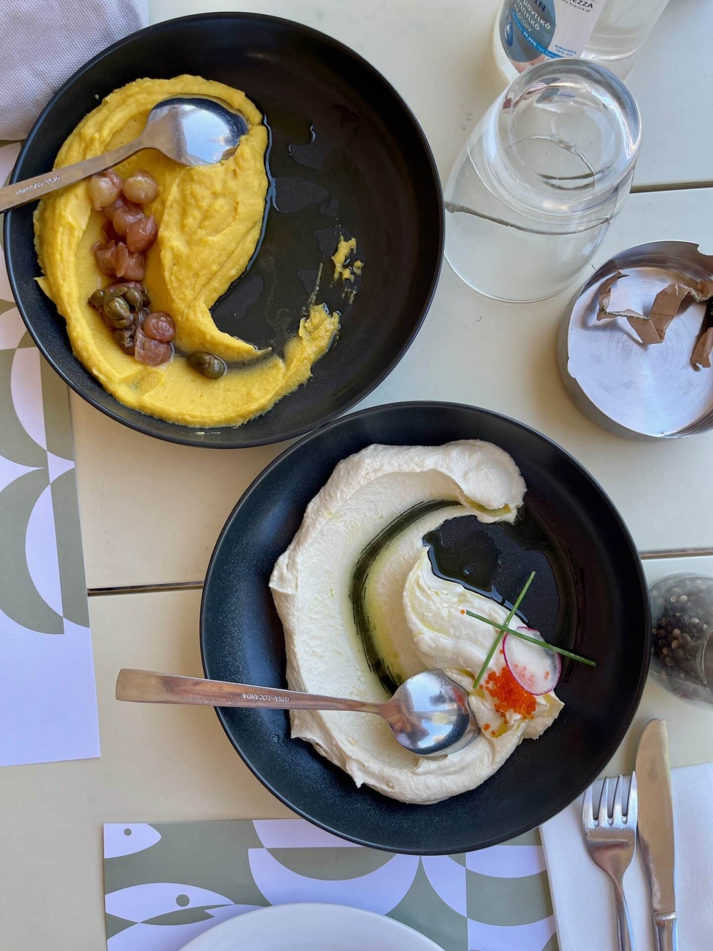 Overhead view of two bowls with creamy dips, one garnished with olives and the other with a drizzle of olive oil and a sprig, on a table with modern decor.