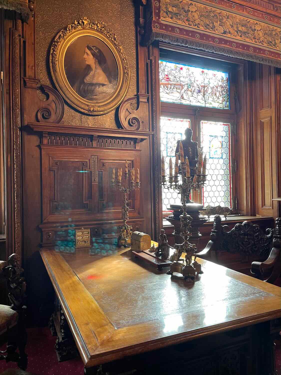 Richly carved wooden interior of a castle with an ornate portrait, detailed wall panels, and a stained glass window casting colorful light on an armor suit, exuding historical opulence.