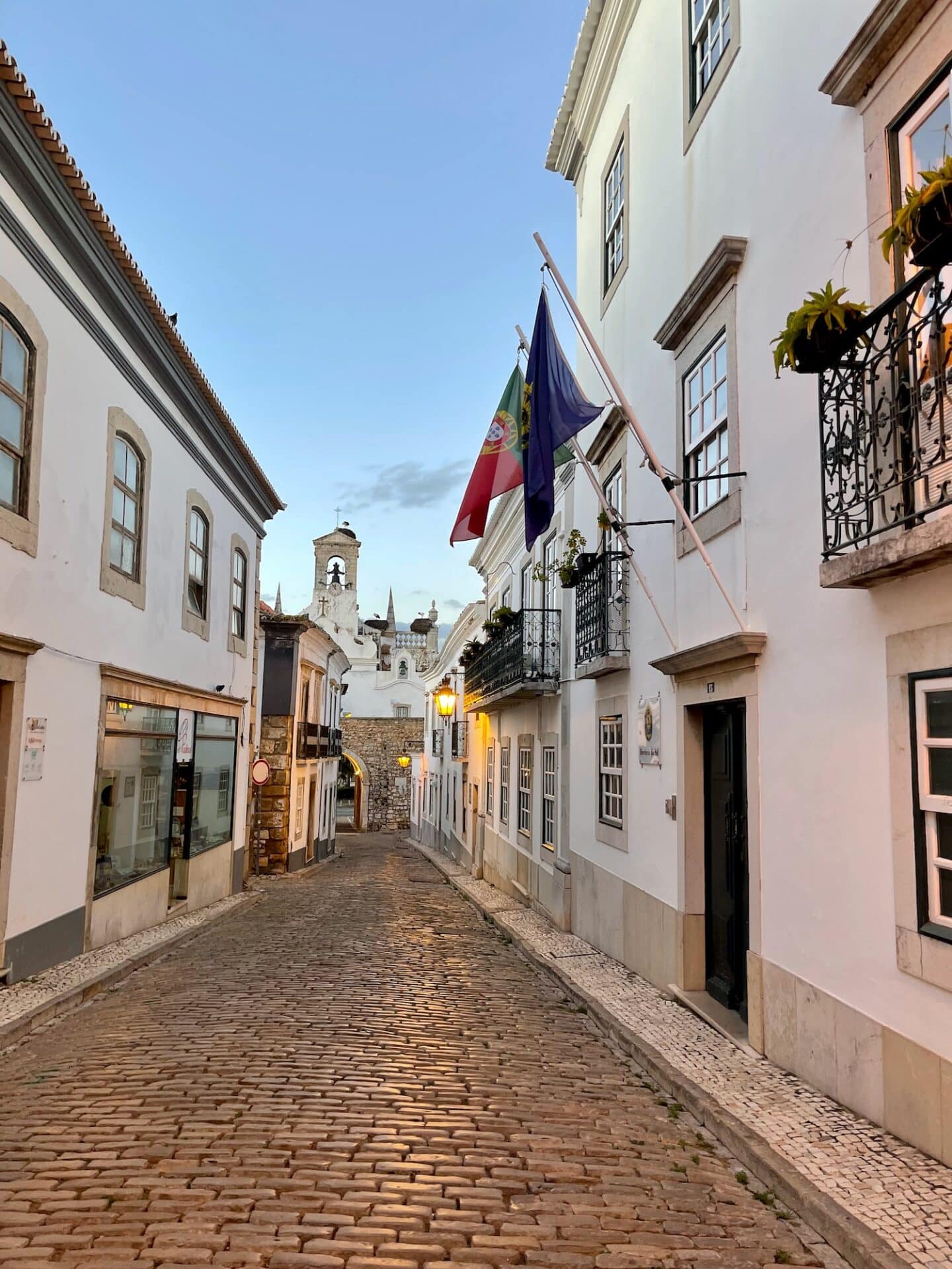 Twilight descends on the cobblestone streets of Faro, with Portuguese and European Union flags hanging above, and a white baroque church in the distance.