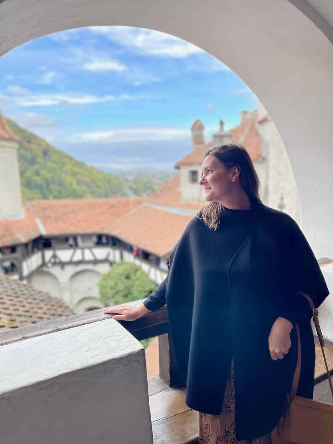 A woman smiles, looking out from an arched opening of Bran Castle, with a view of terracotta rooftops below and a serene mountainous landscape in the distance.