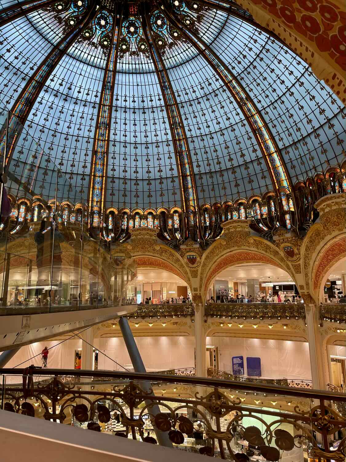 A breathtaking view inside Galeries Lafayette, showcasing its famed stained glass dome and opulent balconies. The golden decor and intricate details create a luxurious shopping environment, highlighted by the warm lighting and the bustling activity below.