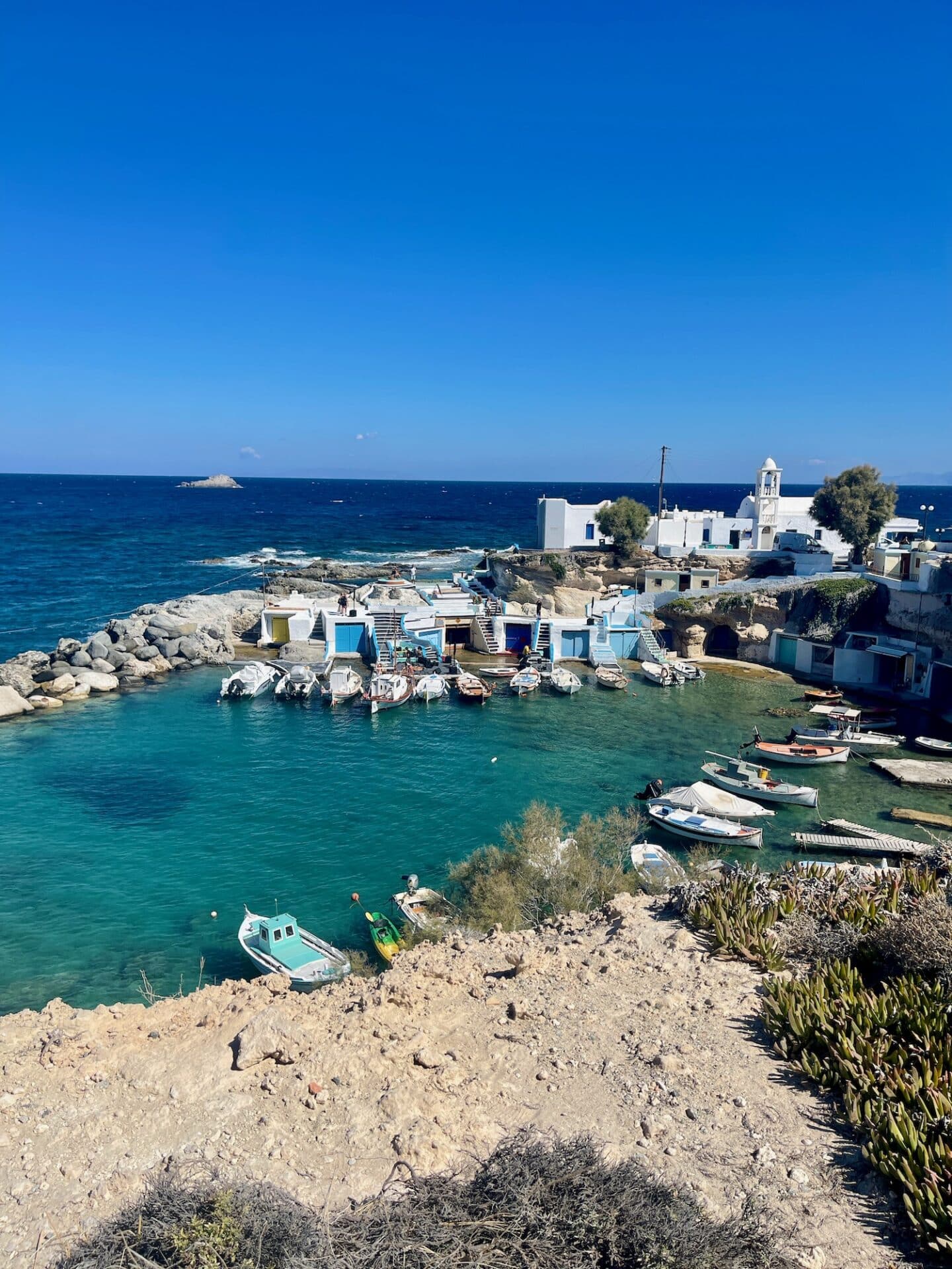 Quaint coastal village with traditional white buildings and a small boat harbor, set against a vivid blue sea and clear sky.