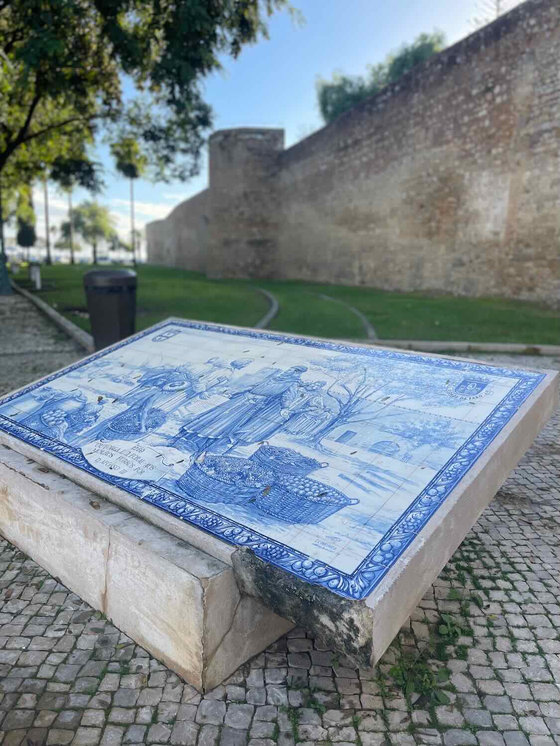 An ornate blue and white ceramic tile mural depicting historical scenes, set on a stone bench with Faro's ancient city wall in the background and a clear blue sky overhead.