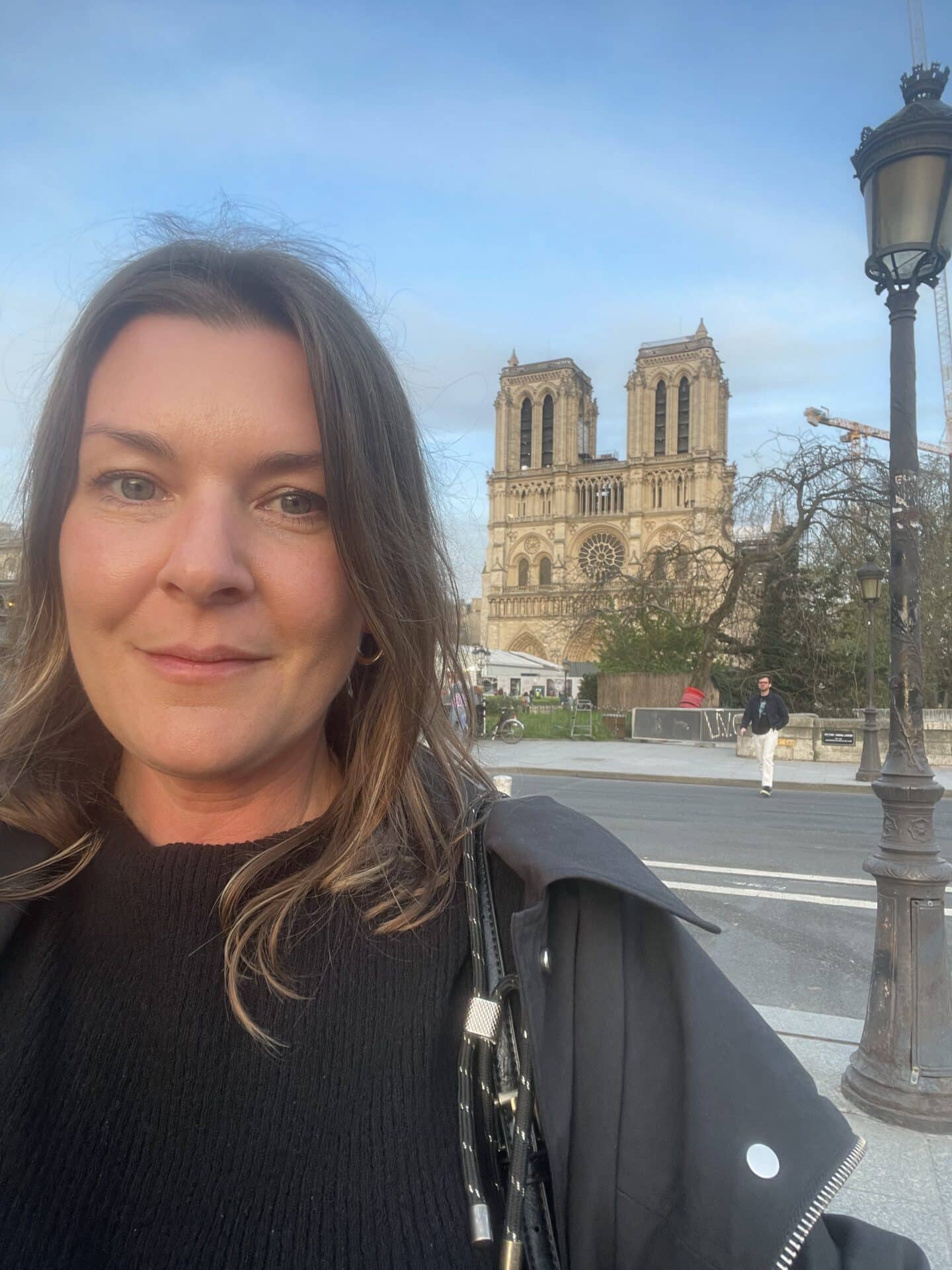 A close-up selfie of a solo female traveler, with the historic Notre Dame Cathedral in the background. Her warm smile and casual style contrast beautifully against the intricate Gothic architecture of the cathedral.