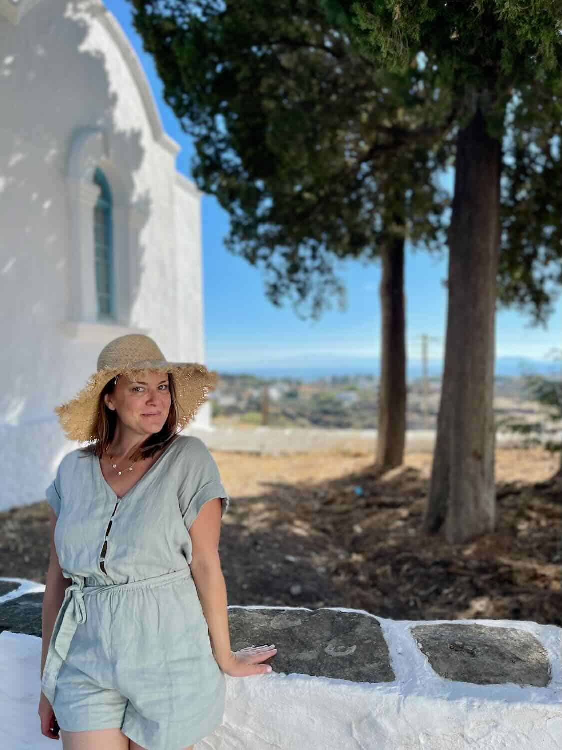A traveler in casual summer attire poses with a straw hat against the backdrop of a traditional white-walled Sifnos church,