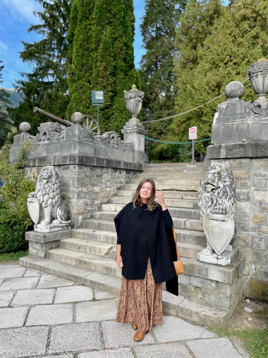 A woman in a flowing skirt and black cape stands at the ornate stone staircase entrance of Peles Castle, flanked by classical statues and vibrant green foliage.