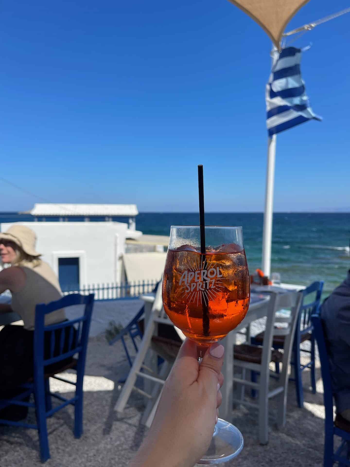 A person holding a glass of Aperol Spritz with a straw, in focus against a seaside taverna backdrop with a fluttering Greek flag, evoking the casual dining atmosphere in Milos.