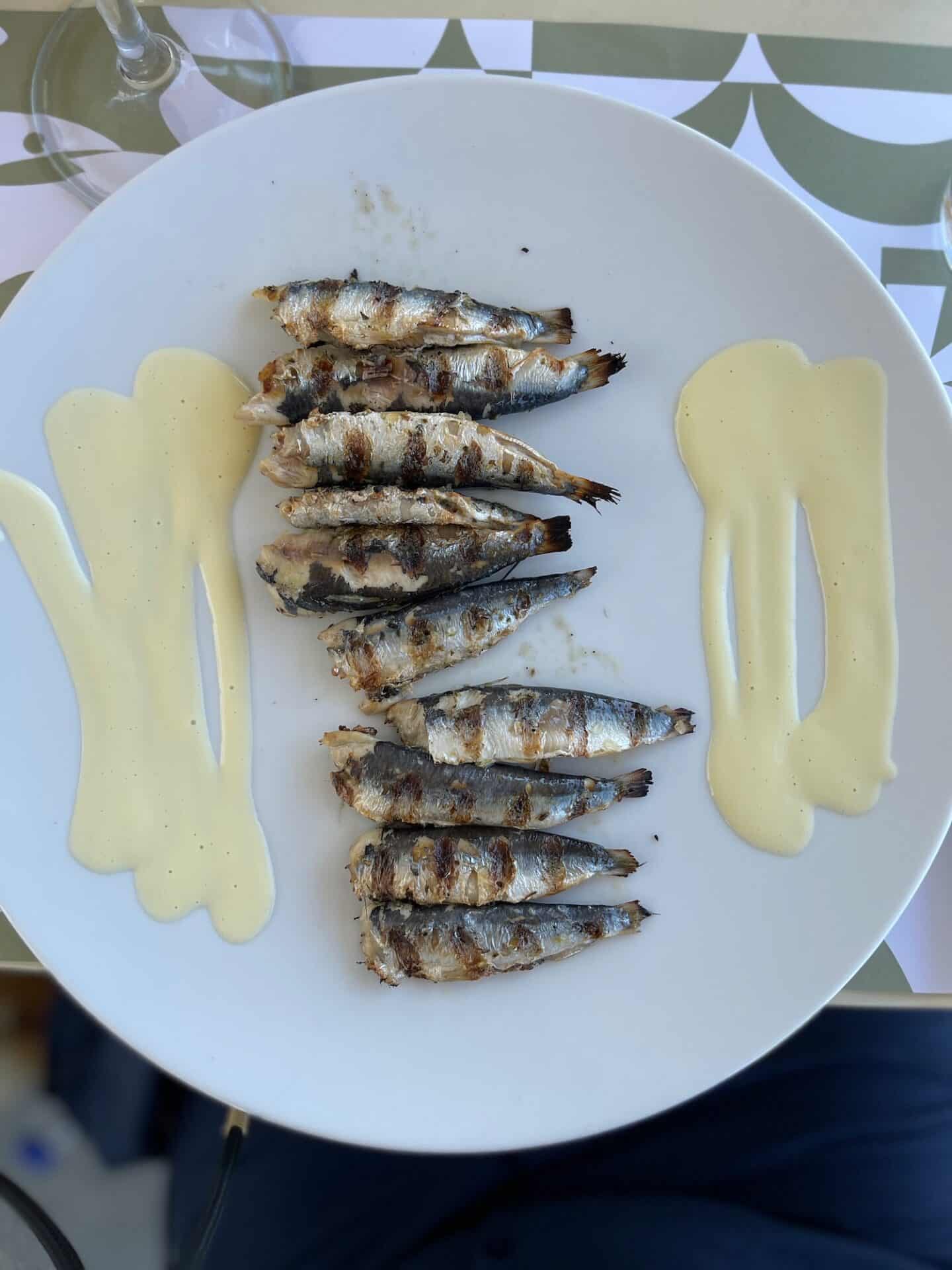 Grilled sardines served on a white plate with a drizzle of yellow sauce on the side, a traditional dish in Milos.