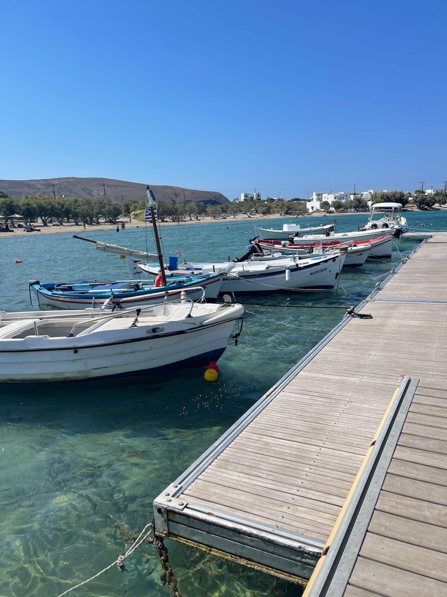Several small boats moored at a wooden pier in Milos, with clear turquoise waters and white buildings in the background under a clear blue sky