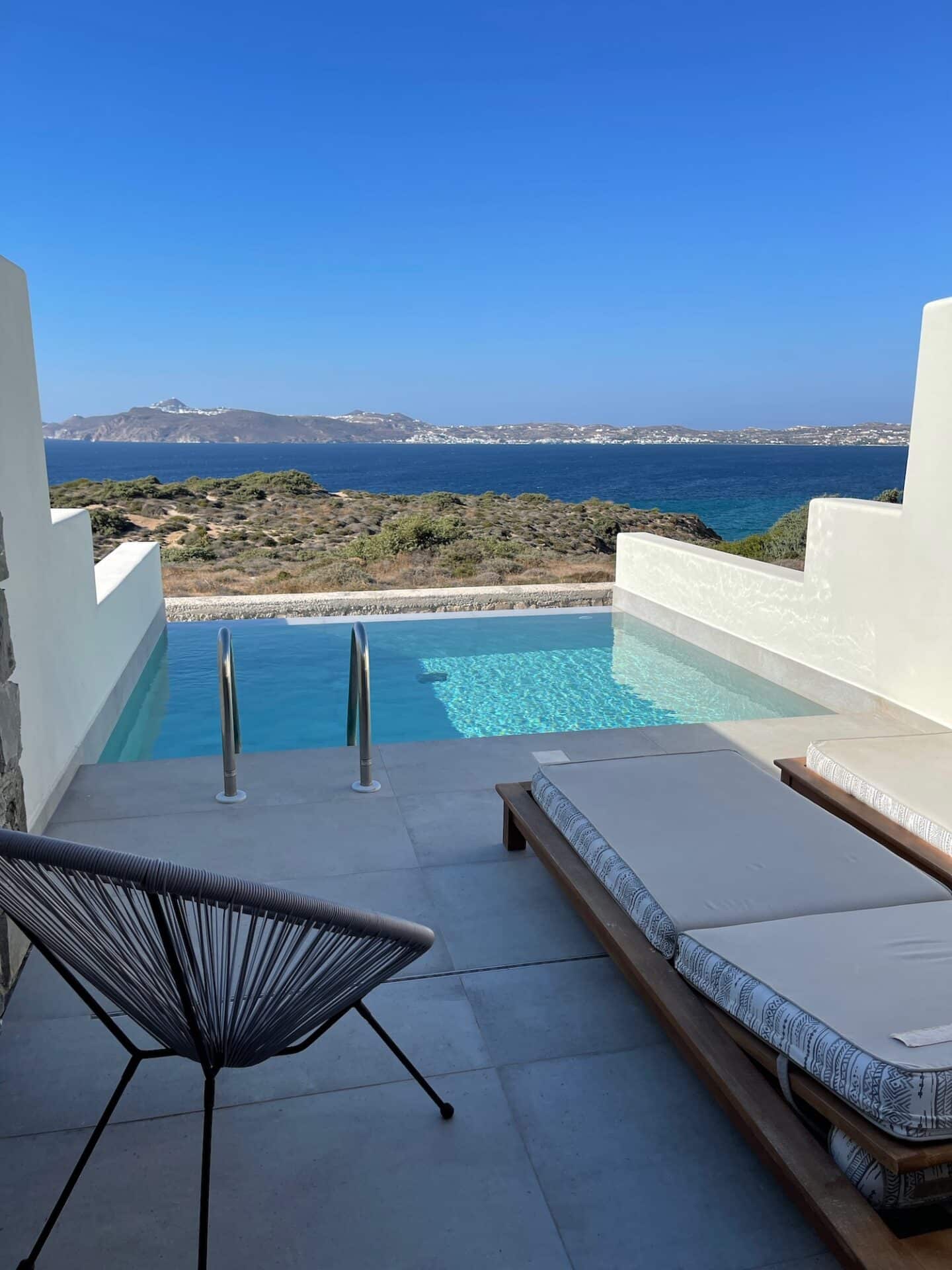 A tranquil infinity pool overlooking the azure Aegean Sea in Milos, flanked by sunbeds and a stylish black wire chair, capturing the essence of luxury Greek island living.