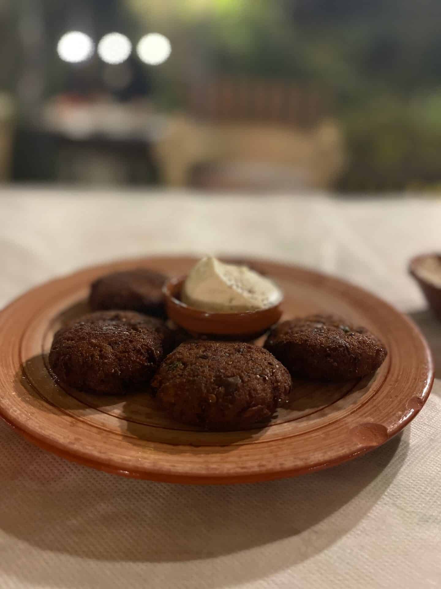 Close-up of crispy fried falafel served on a terracotta plate with a side of creamy sauce, presented on a table with soft, ambient lighting.
