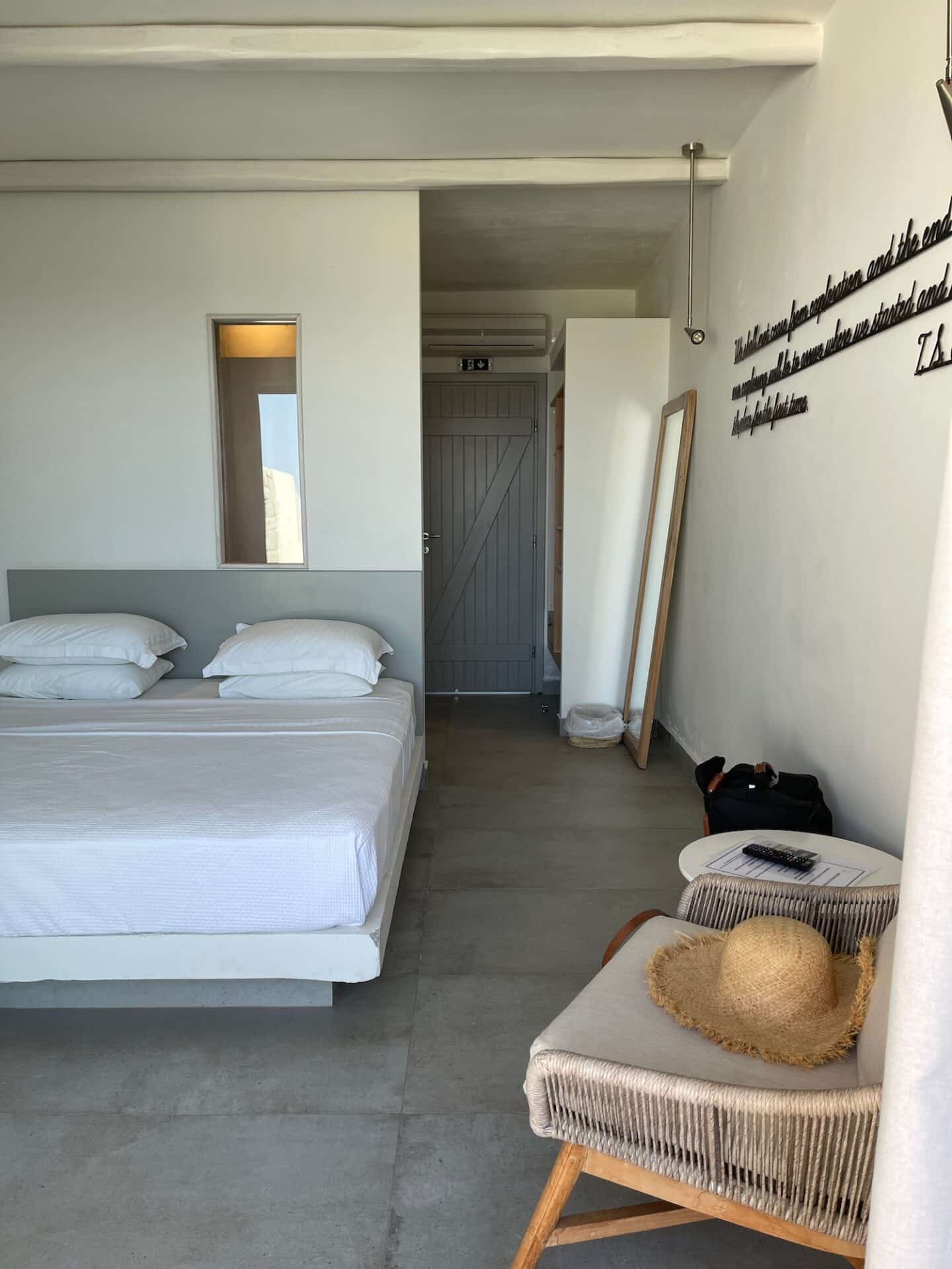 Minimalist style bedroom in Milos with two single beds, a woven hat on a chair suggesting a relaxed holiday vibe, and a mirrored door reflecting the bright, airy space.