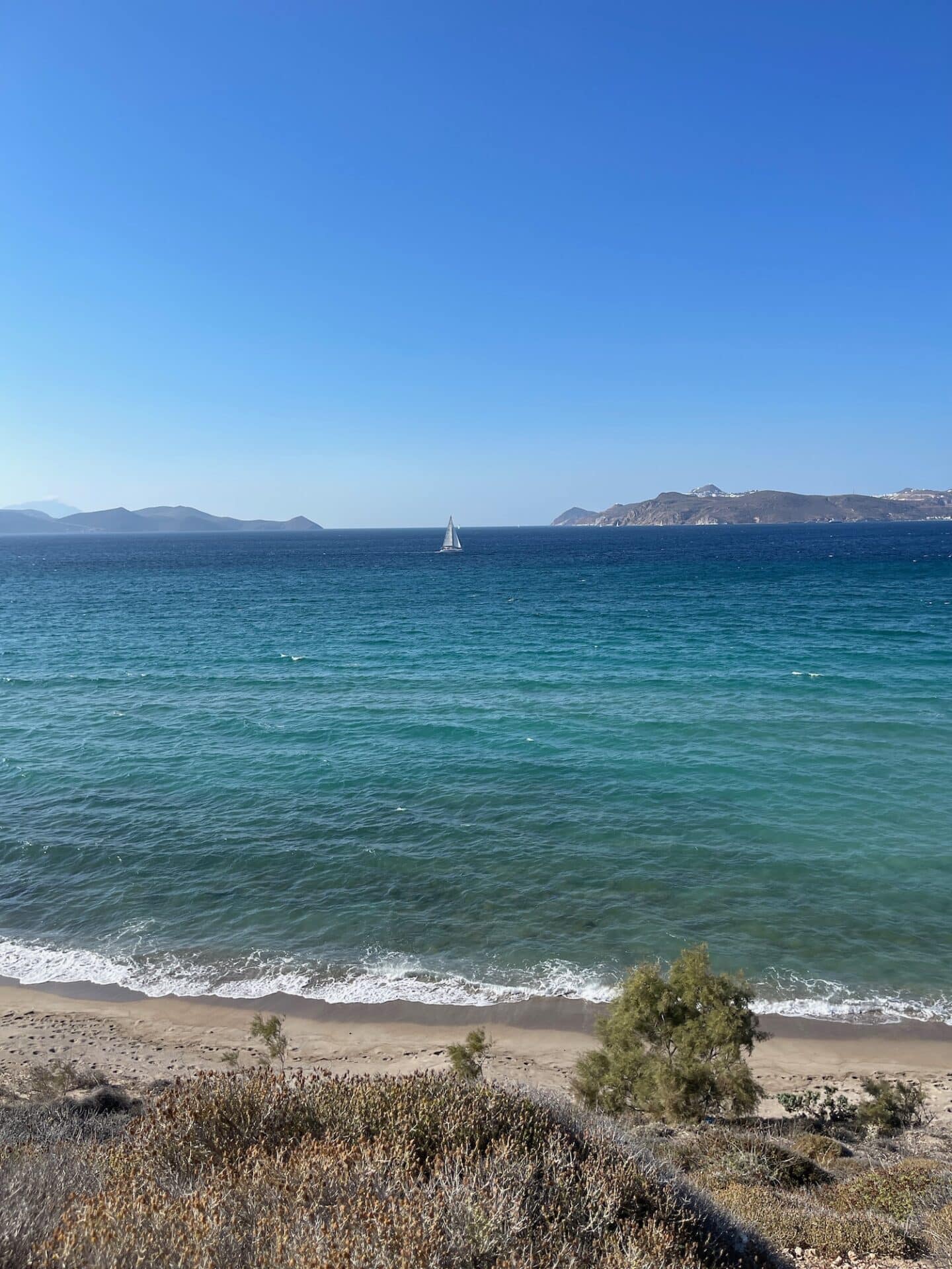 A scenic view of a sandy beach in Milos, overlooking a calm sea with a single sailboat in the distance, and mountainous terrain under a clear blue sky.