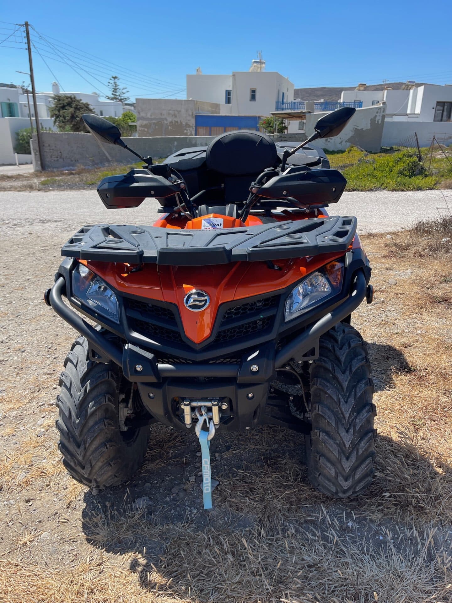 Front view of an orange ATV quad bike with a 'CFMOTO' logo, ready for an adventurous ride around the rugged terrain of Milos, with white Cycladic houses in the background.
