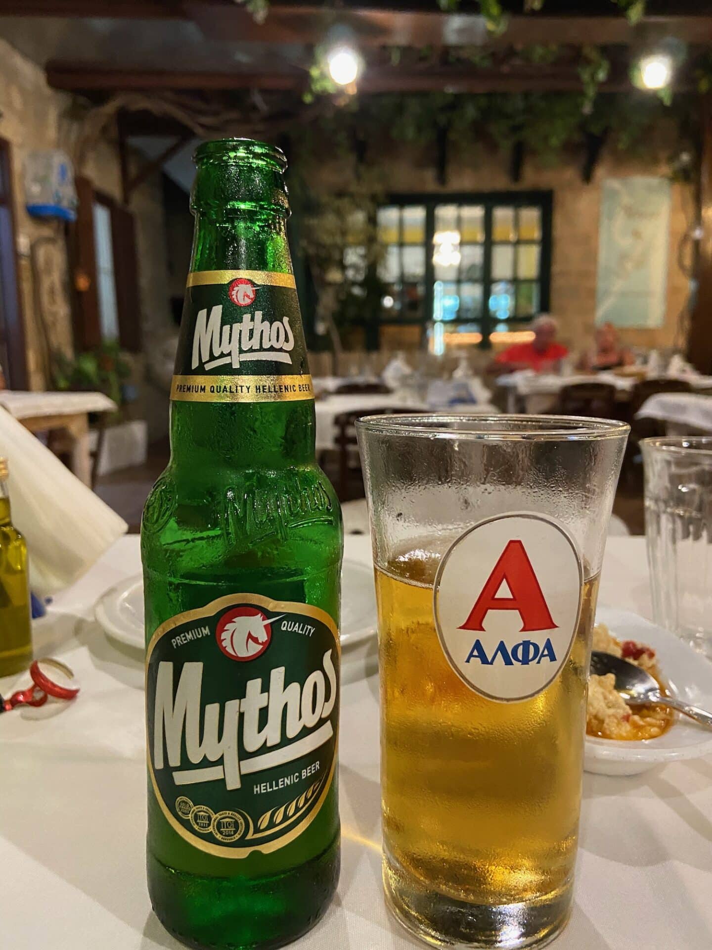A close-up of a cold Mythos beer bottle beside a glass half-filled with beer, with a blurry taverna scene in the background, capturing the essence of a relaxed evening in Milos.