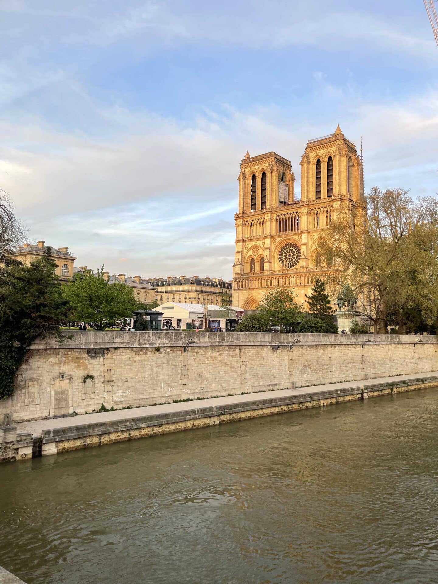A serene image capturing Notre Dame Cathedral at sunset. The golden hue of the setting sun highlights the intricate facade and twin bell towers of the cathedral, contrasting beautifully with the soft blue sky. The Seine River in the foreground reflects the warm colors, enhancing the peaceful yet majestic atmosphere of this historic landmark.