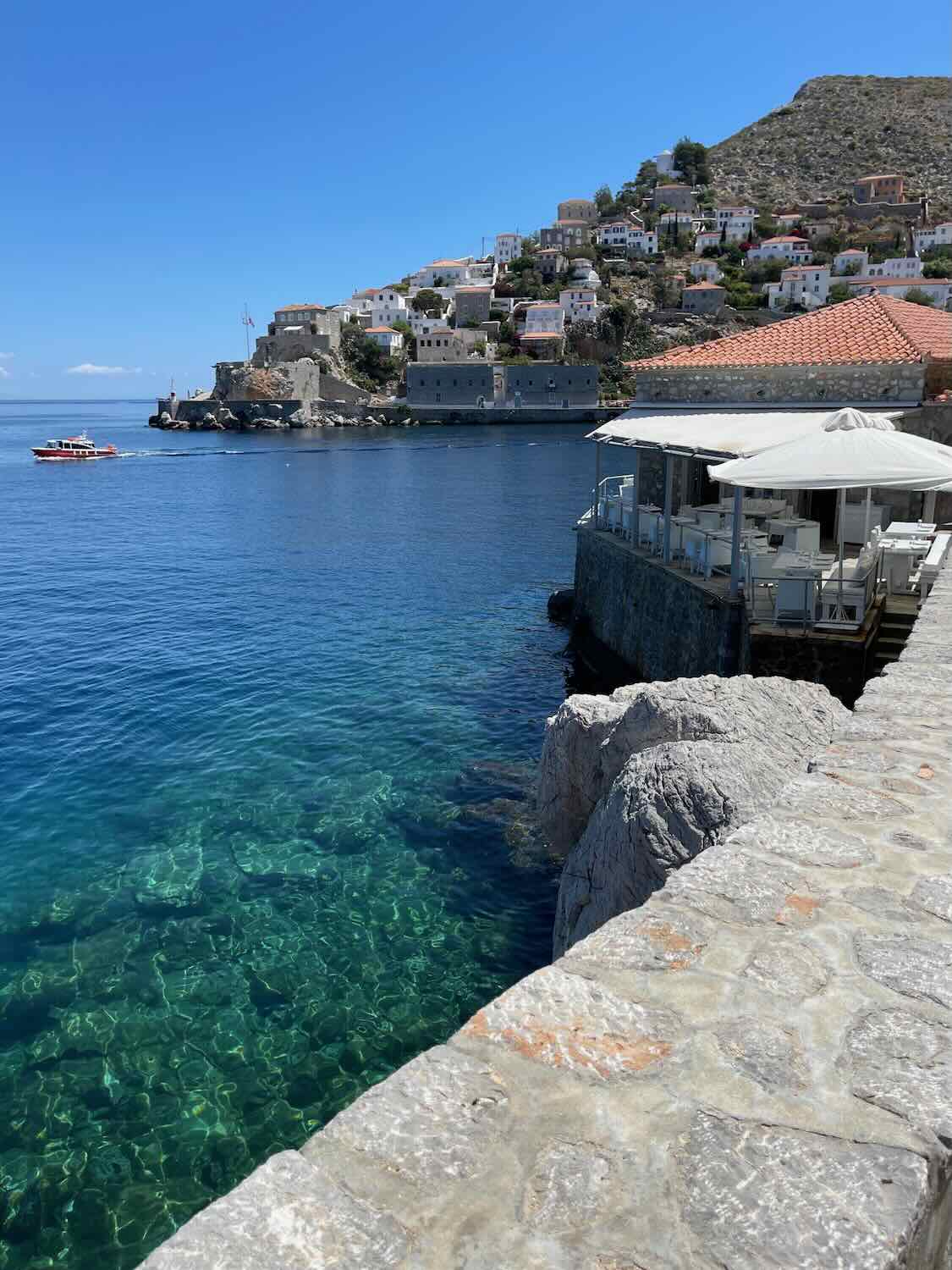 Scenic view of the waterfront and terraced houses of Hydra from Omilos, showcasing the serene blue waters and clear skies on a sunny day.
