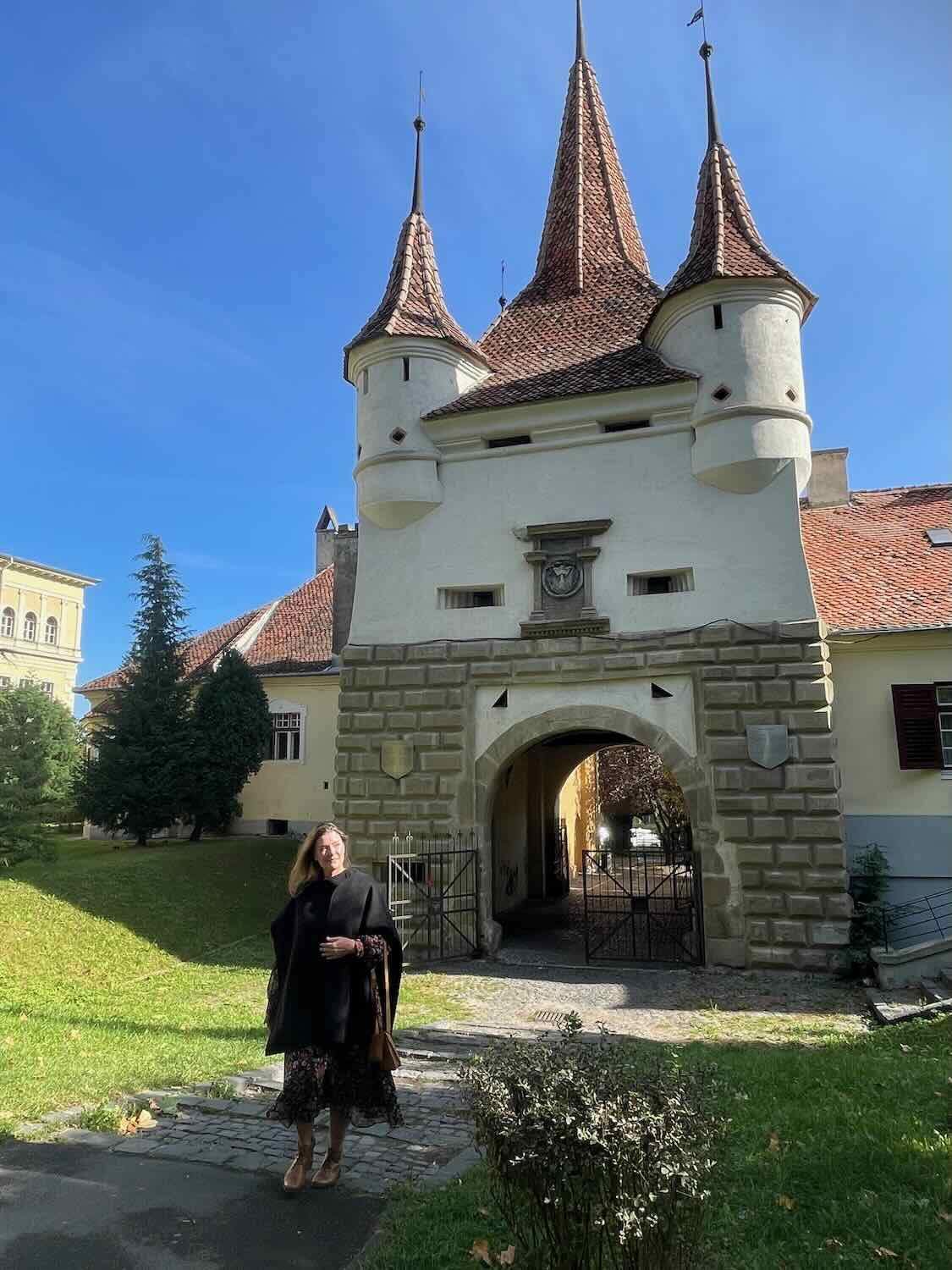 The woman walks away from a castle gateway, dressed in a cape, suggesting a narrative of exploration and departure amidst historical Romanian architecture.