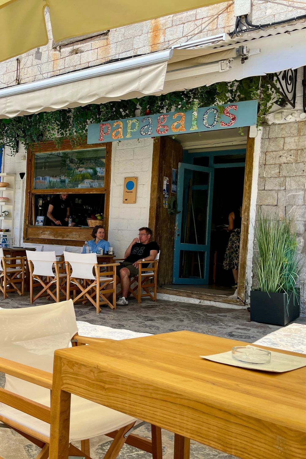 Casual outdoor dining at Papagalos restaurant in Hydra, with patrons enjoying the ambiance under the shade of a vine-covered awning, epitomizing the island's relaxed dining culture.