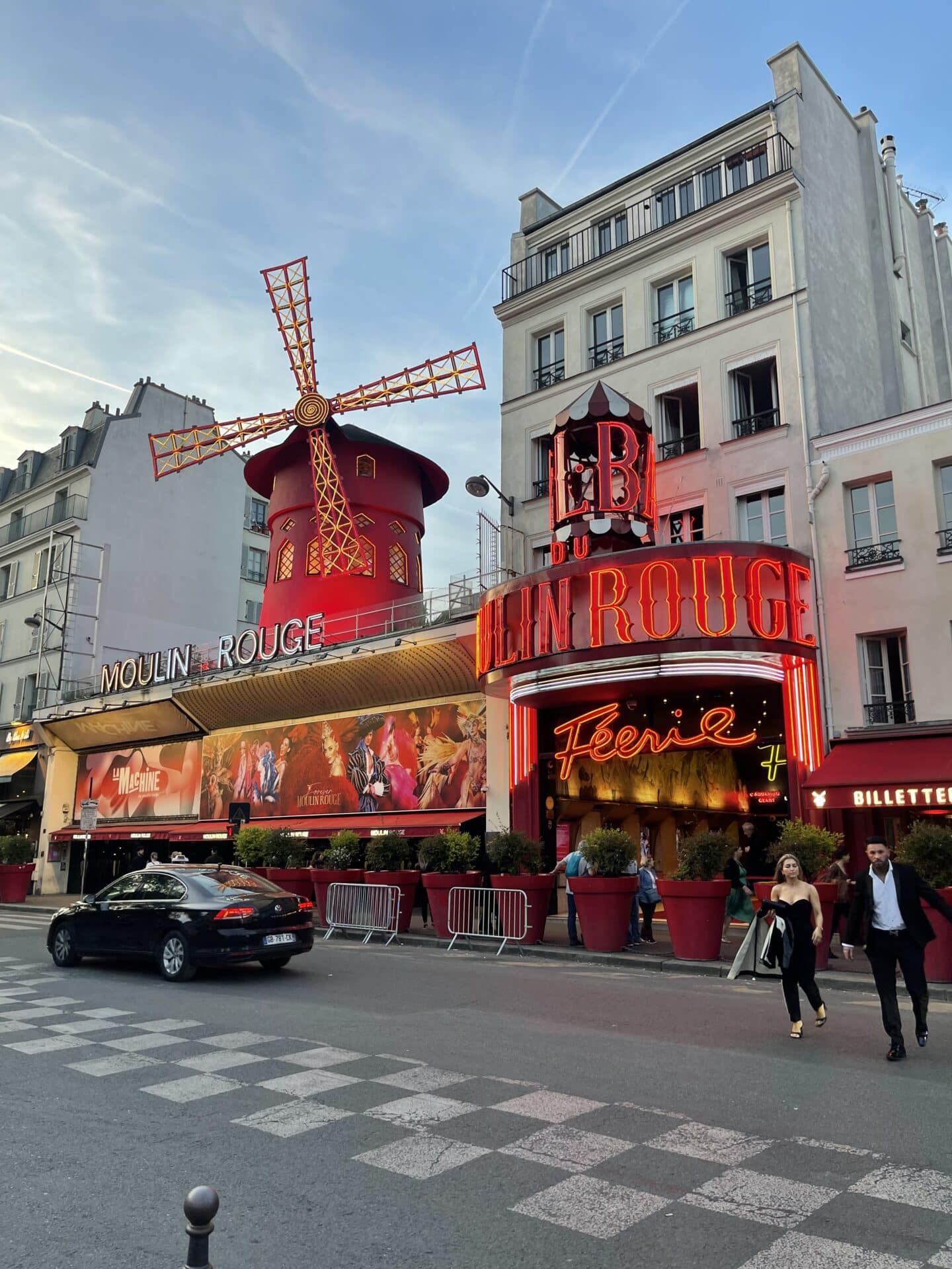 The iconic Moulin Rouge lit up at dusk, showcasing its famous red windmill against a twilight sky, with people walking and cars driving by, capturing the vibrant nightlife of Paris.