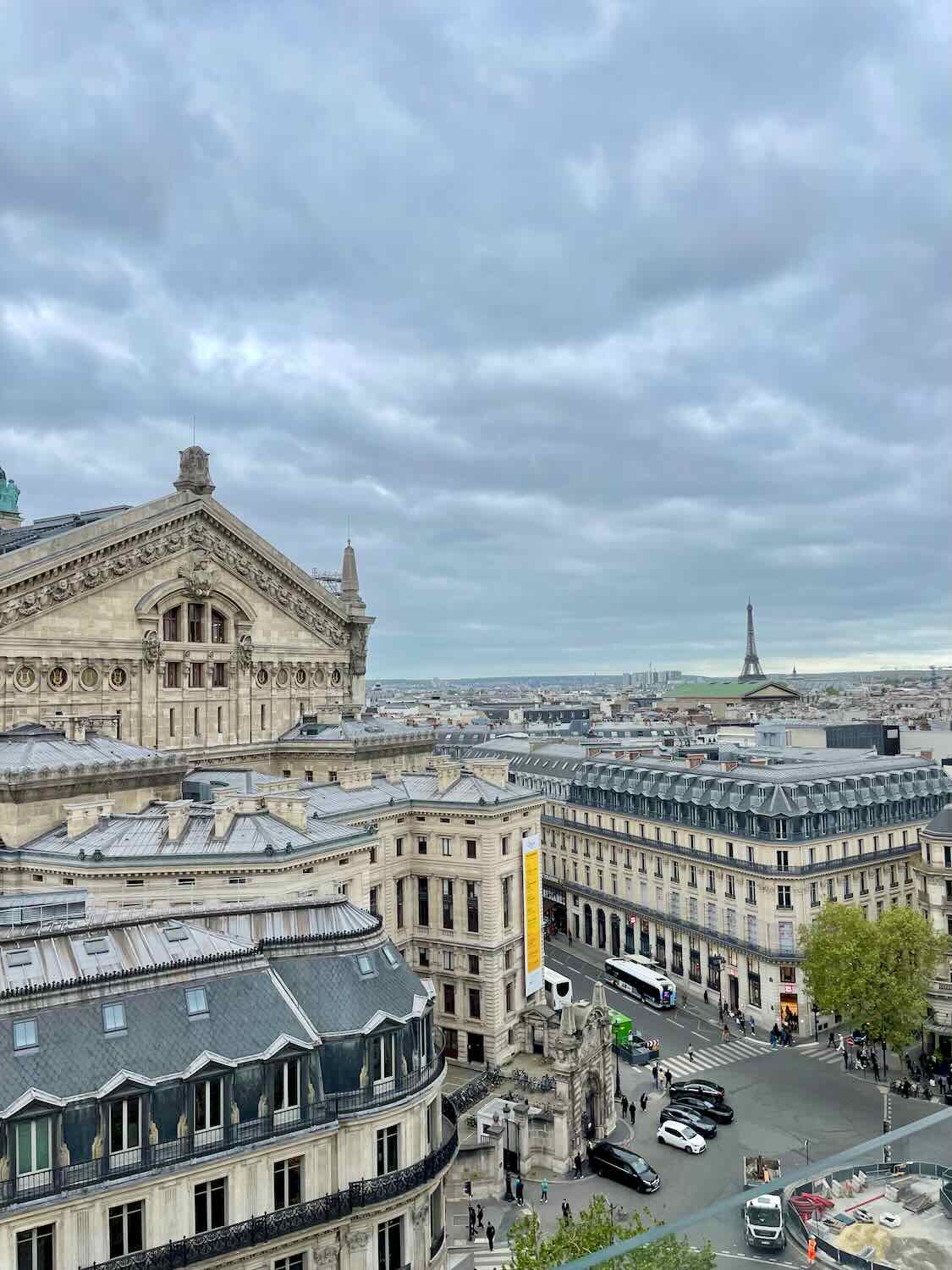 A sweeping view from a rooftop in Paris, overlooking iconic cityscape elements including grand buildings and distant views of the Eiffel Tower, set against a dramatic cloudy sky. The scene captures the vast, dynamic architecture of Paris from an elevated perspective.