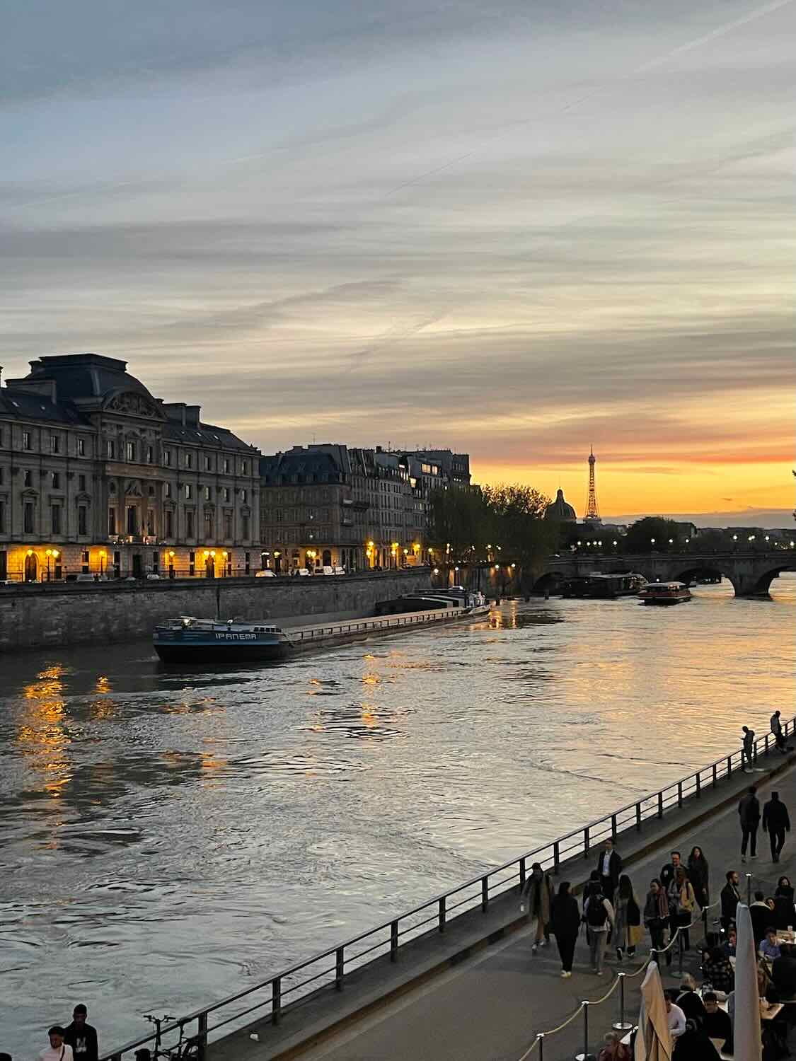 A breathtaking evening view of Paris from a riverside perspective. The sunset casts a warm glow over the city, illuminating the Seine River and the silhouettes of historical buildings. The Eiffel Tower in the distance is lit up, providing a striking contrast against the twilight sky, capturing the romantic essence of Paris at night.