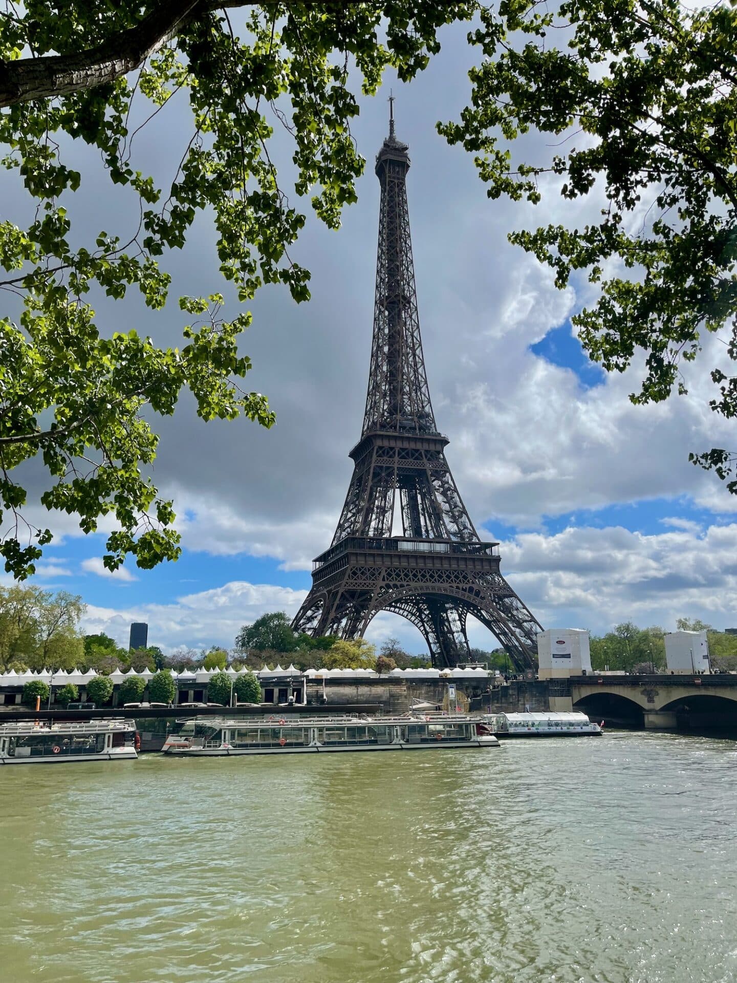 The Eiffel Tower stands majestically against a cloudy sky, viewed from the riverbank with verdant tree branches arching overhead. A river cruise boat is docked along the Seine, waiting for passengers to embark on a scenic journey through the heart of Paris.