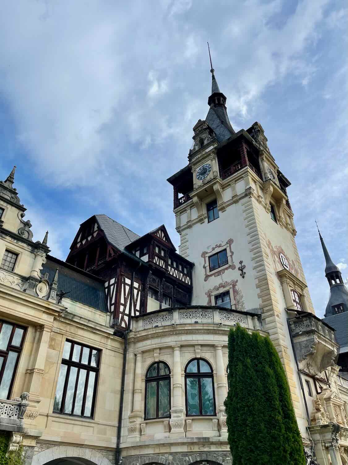 An exterior shot of Peles Castle showcasing its fairy-tale towers and mix of architectural styles, set against a backdrop of clear blue skies and fluffy clouds.