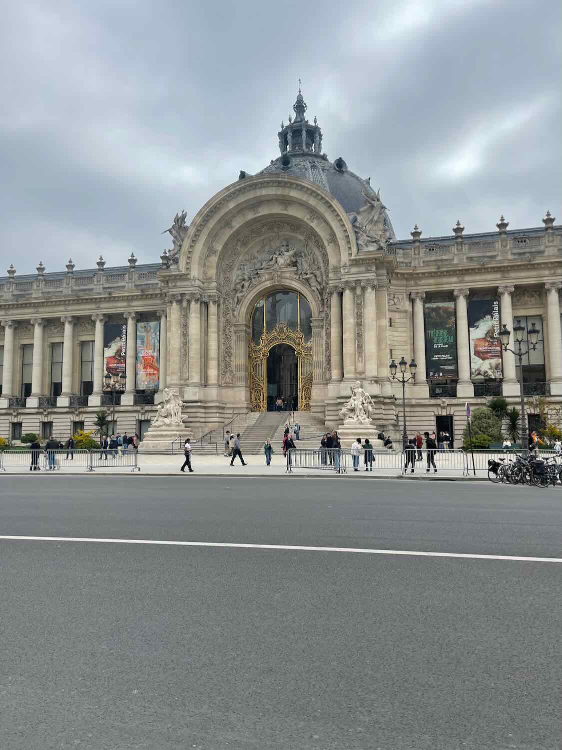 The grand entrance of the Petit Palais, featuring ornate architectural details and sculptures, under a cloudy sky, with pedestrians and cyclists passing by, reflecting the cultural richness of Paris.