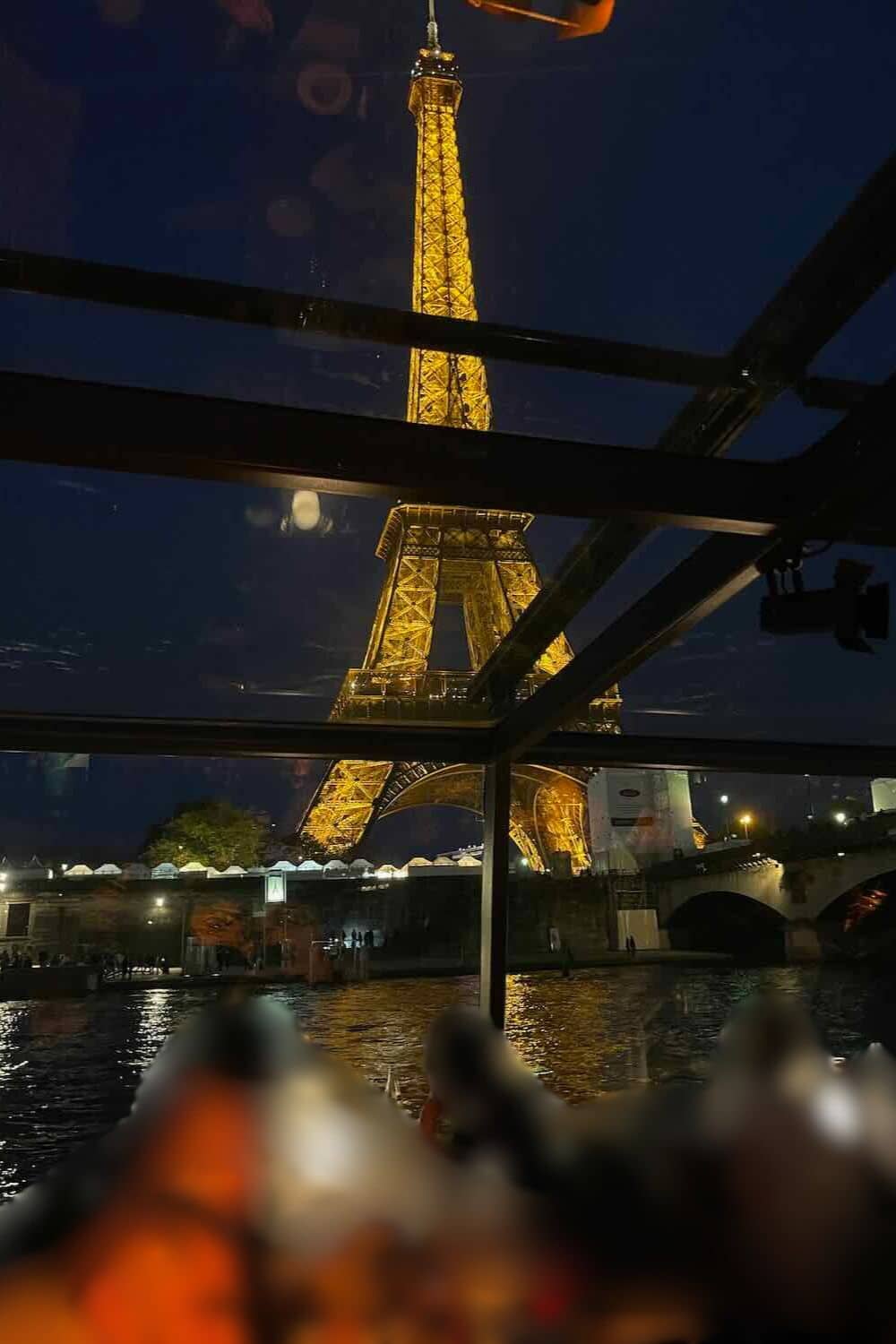 A night scene on a river cruise in Paris, with diners enjoying their meals under the illuminated Eiffel Tower visible through the glass roof, reflecting off the water for a magical dining experience.