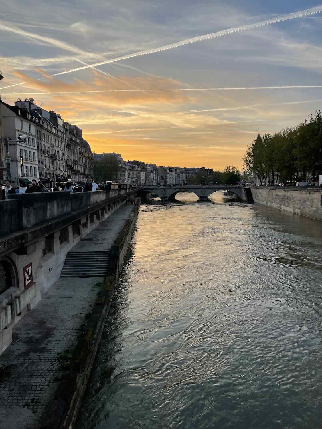 A serene river walk scene in Paris at sunset, showing the Seine River reflecting the orange sky, with historical buildings lining the banks and people enjoying the peaceful evening.