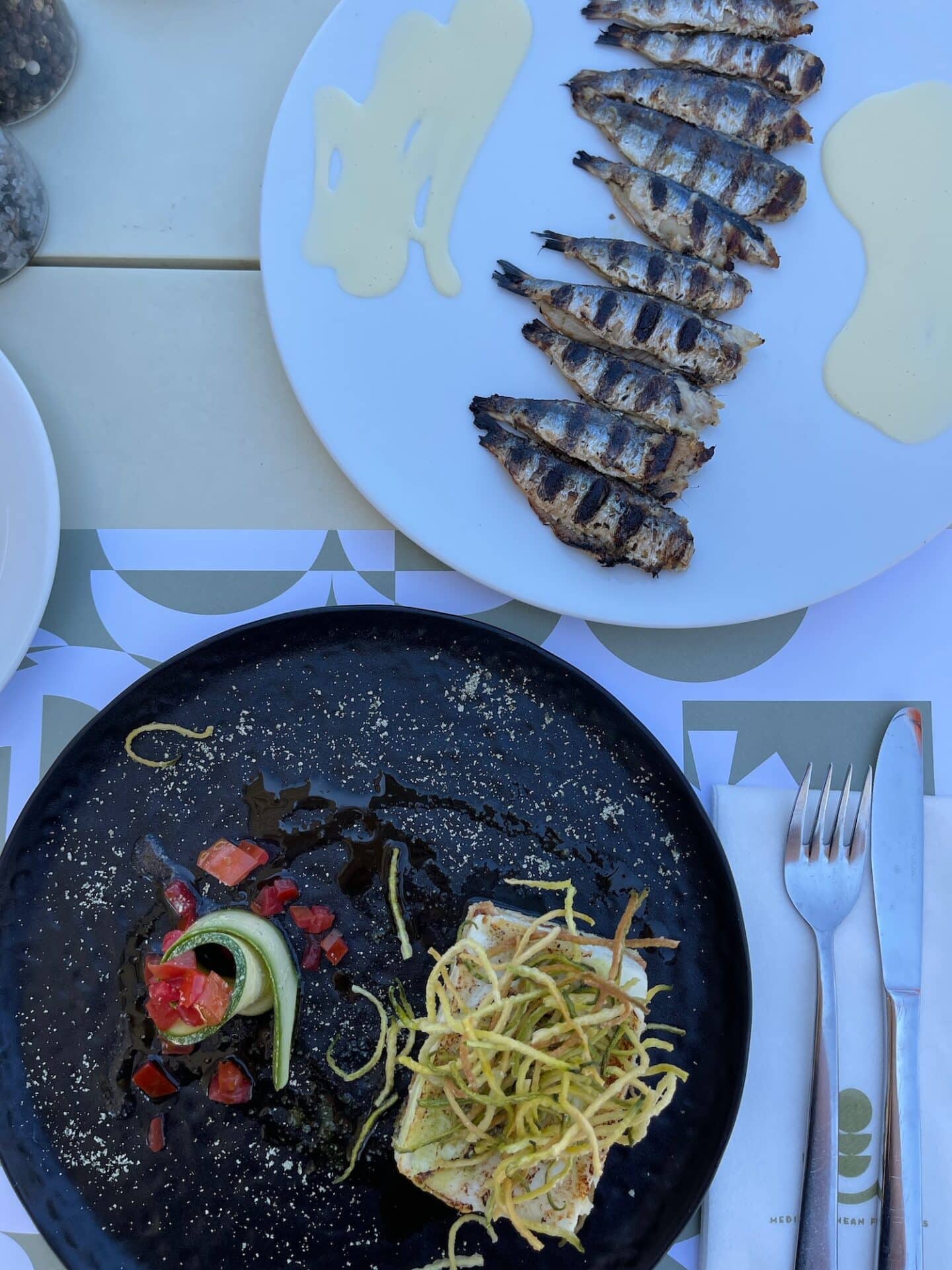 An overhead view of a Greek meal featuring grilled fish on a black plate, pasta, and two artistic splashes of sauce on a white plate, typical cuisine in Milos.