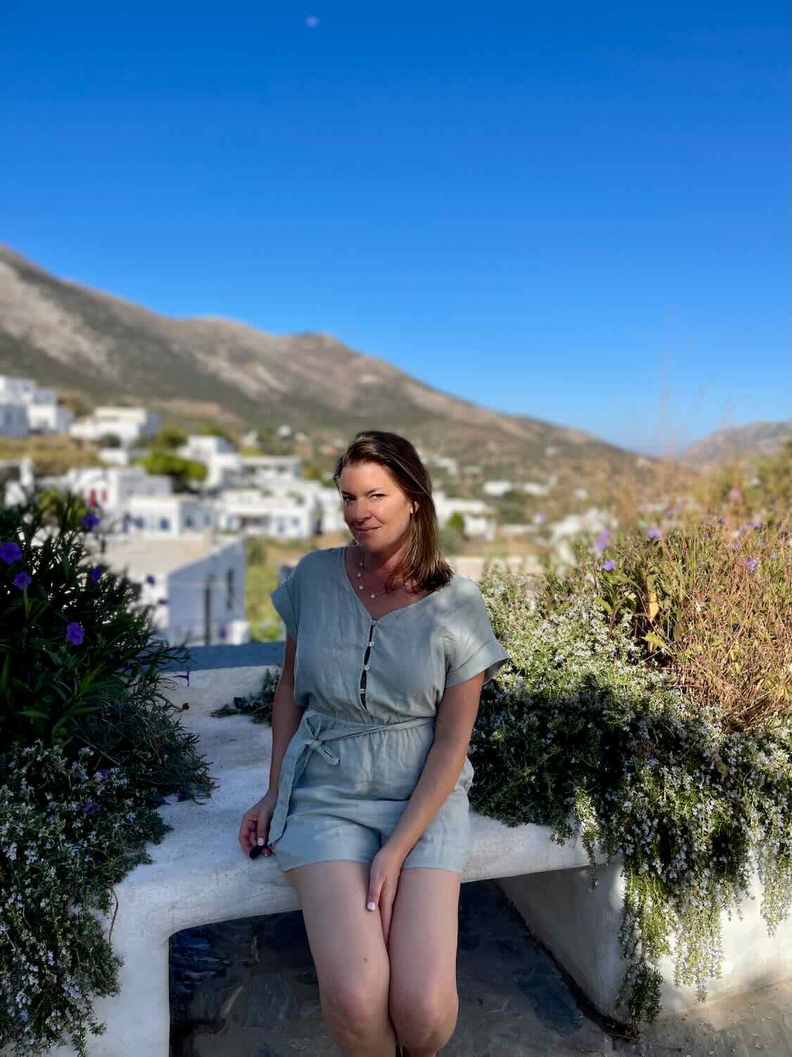 A woman smiles gently while seated on a white bench in Sifnos, with lush greenery and mountains in the background, a serene moment to include in a Sifnos itinerary.