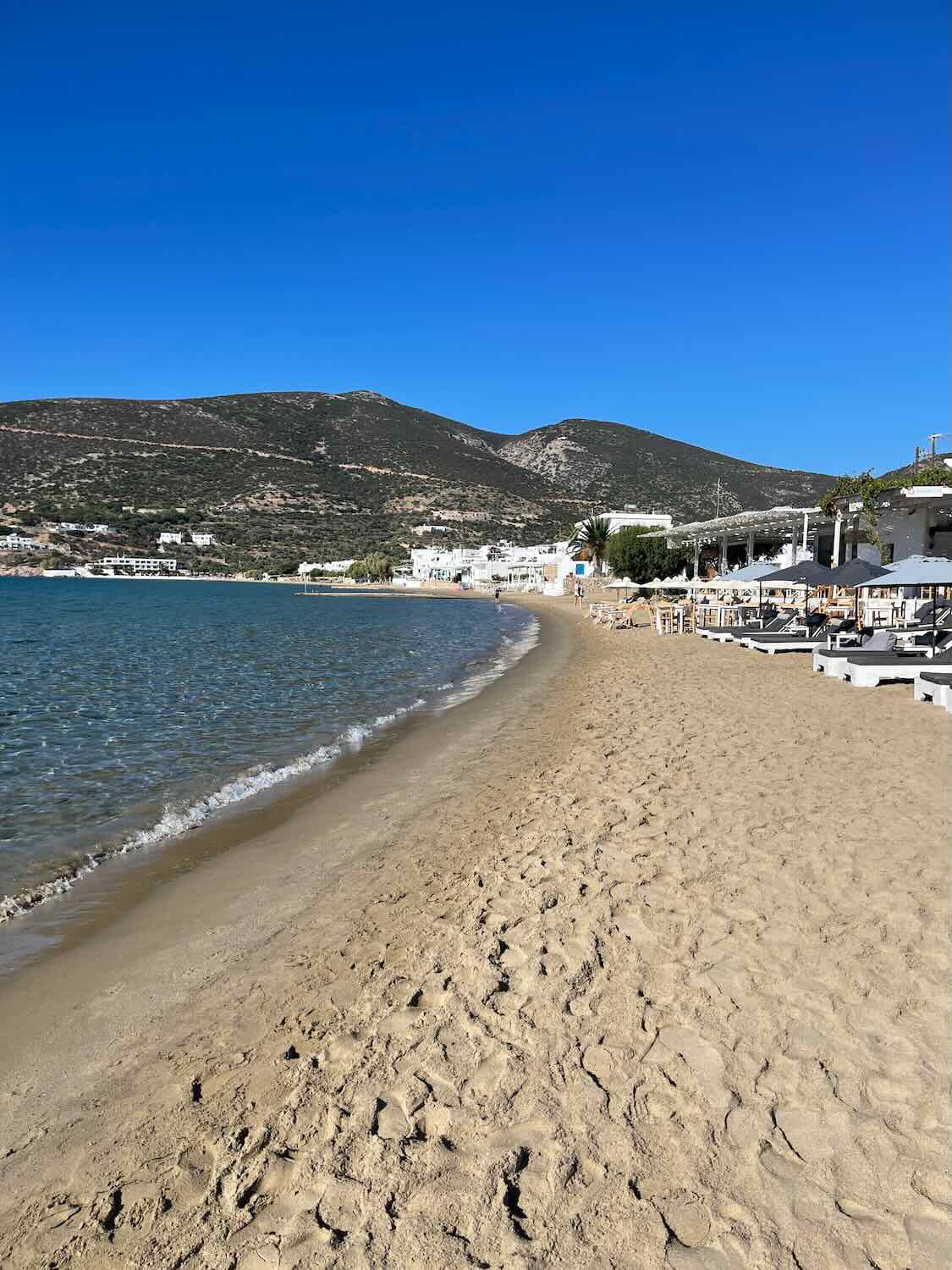 A tranquil beach scene with a long stretch of golden sand curving along the calm blue waters of Sifnos,