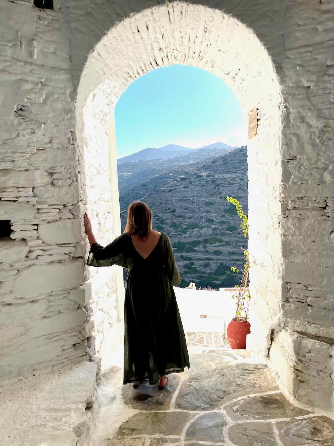 A woman in a flowing dress stands at the arched entrance of Kastro, overlooking the terraced landscape of Sifnos, a historic site to explore on a Sifnos.