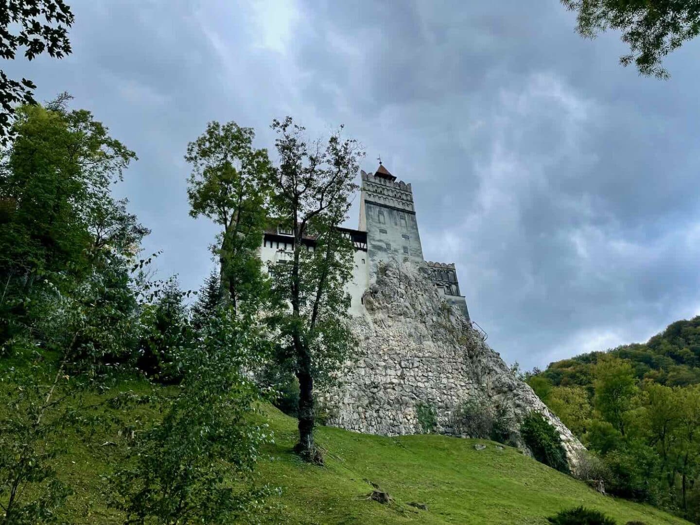 A striking angle of Bran Castle standing atop a steep cliff, surrounded by lush greenery and overshadowed by a moody, cloud-filled sky, enhancing the castle's haunting presence.