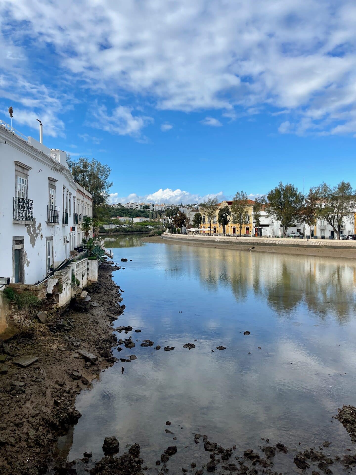 Calm waters of the Gilão River mirror the sky, flanked by the white facades of traditional buildings in Tavira's riverside promenade.