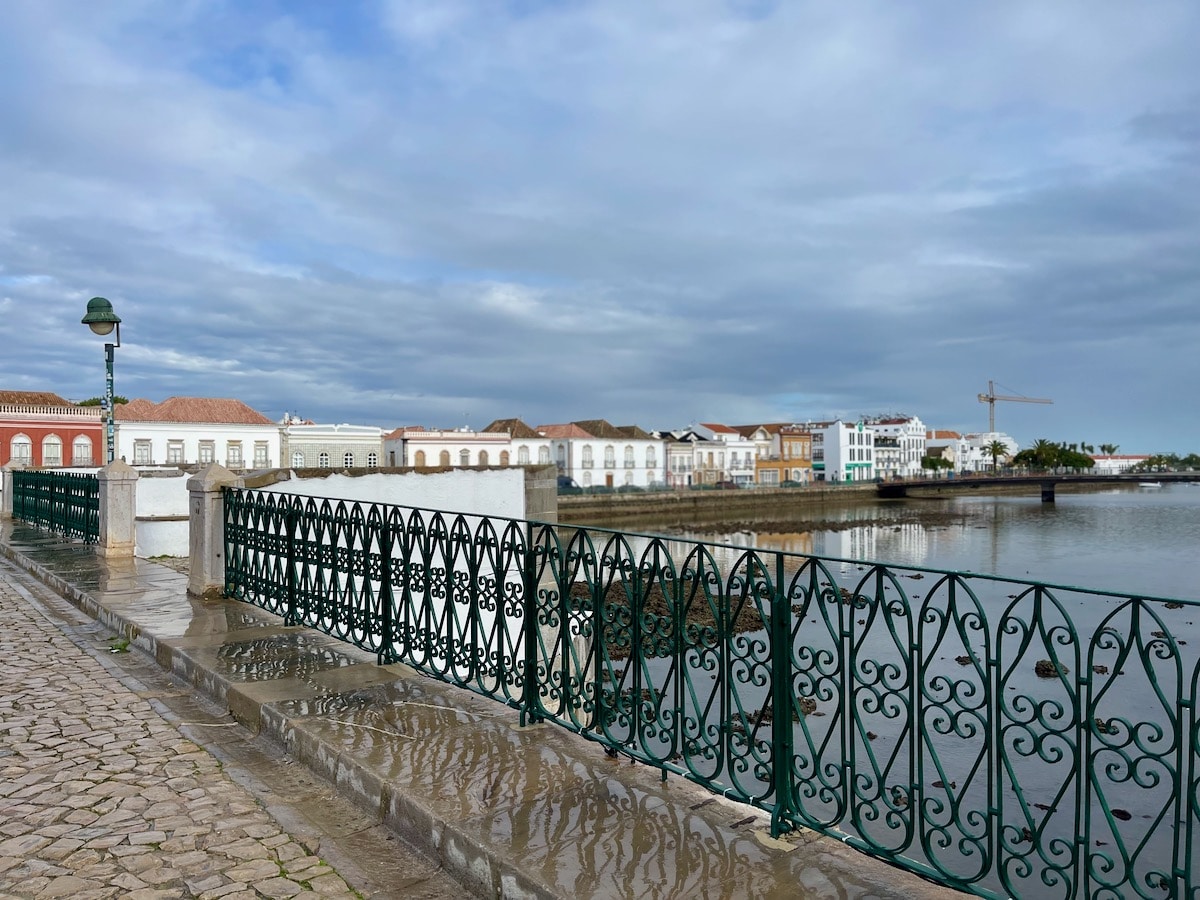 View from a Tavira bridge with ornate green railings, overlooking a serene river and rows of charming white houses, reflecting the peaceful atmosphere of the town.