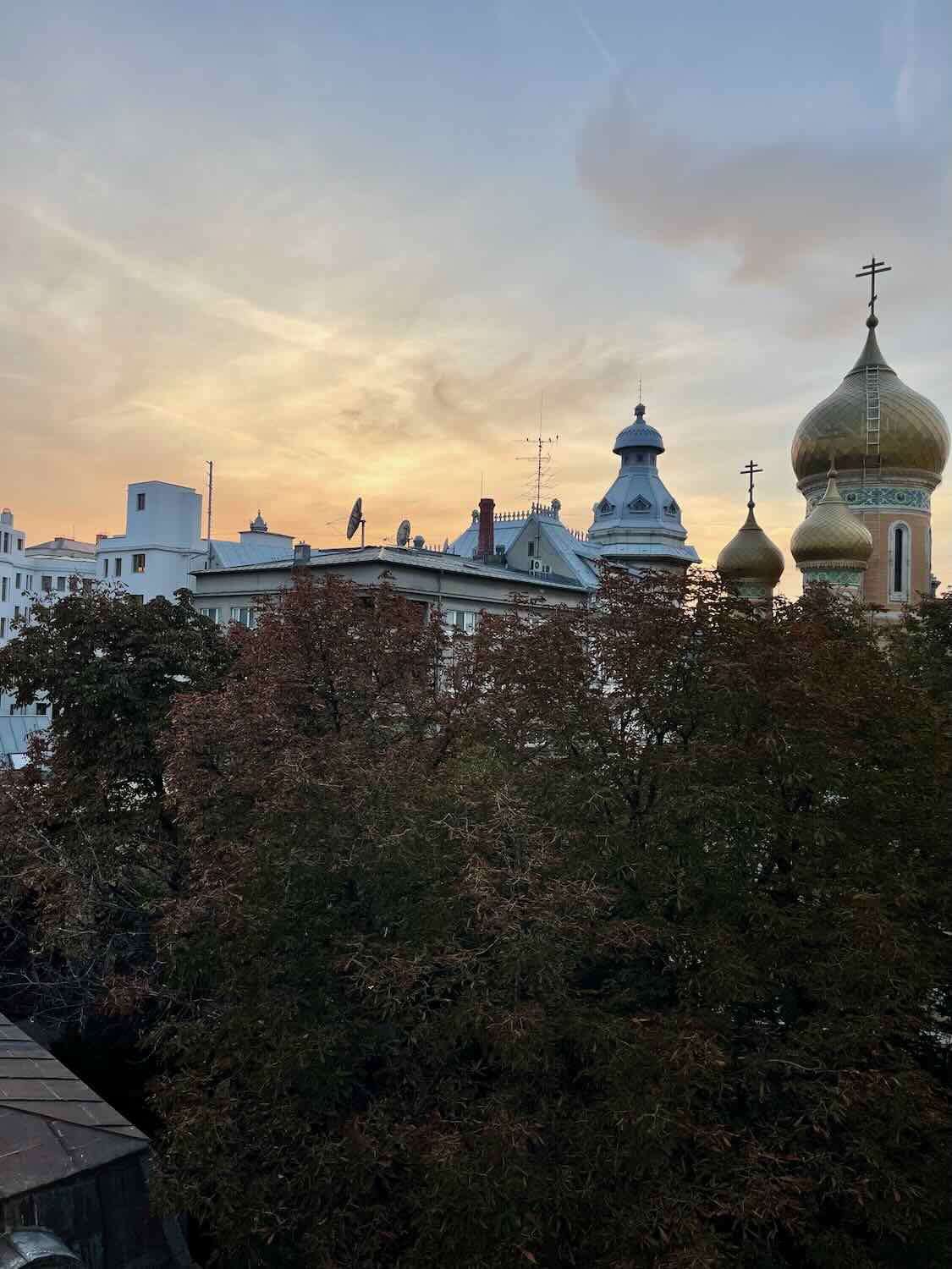 A sunset view over Bucharest, featuring silhouettes of buildings against a sky painted in shades of orange and blue, with the domes of a church adding a distinctive skyline.