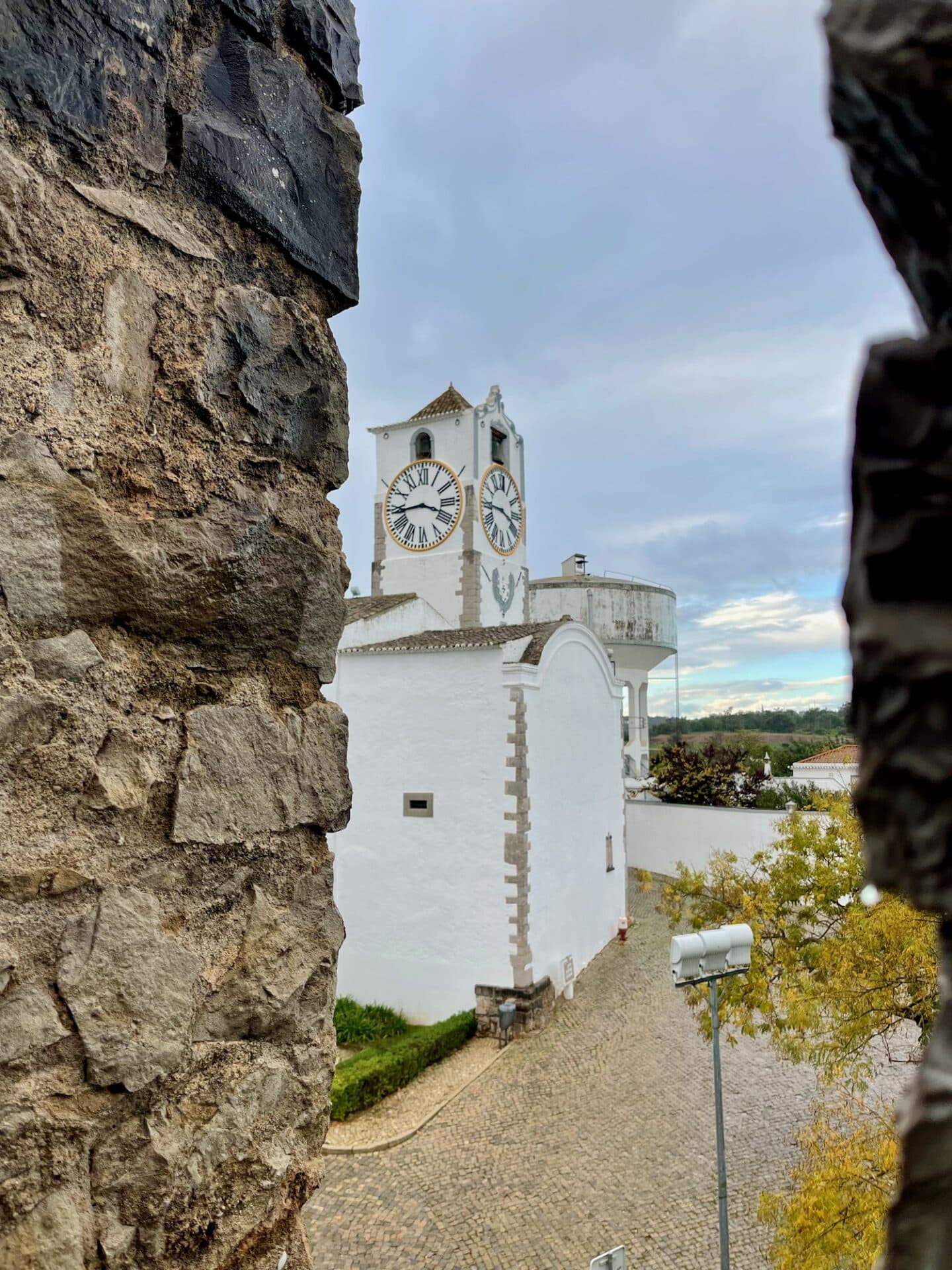 A historic white clock tower seen through the battlements of a castle wall, blending the old architecture with a view of modern street lamps in Tavira.