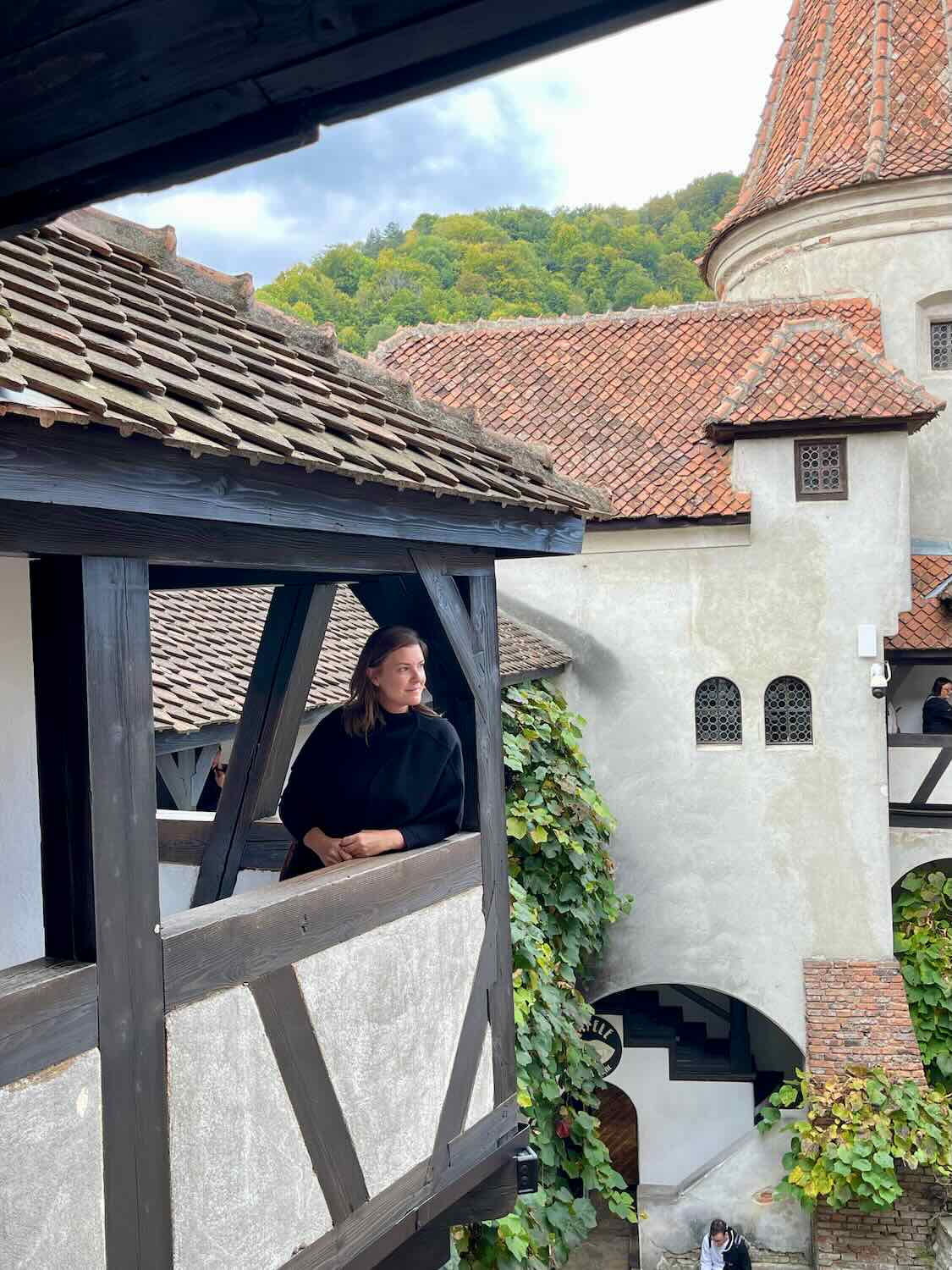 A woman gazes thoughtfully from a wooden balcony at Bran Castle, with the ancient rooftops and dense green forests in the background, offering a personal perspective of the historic site.