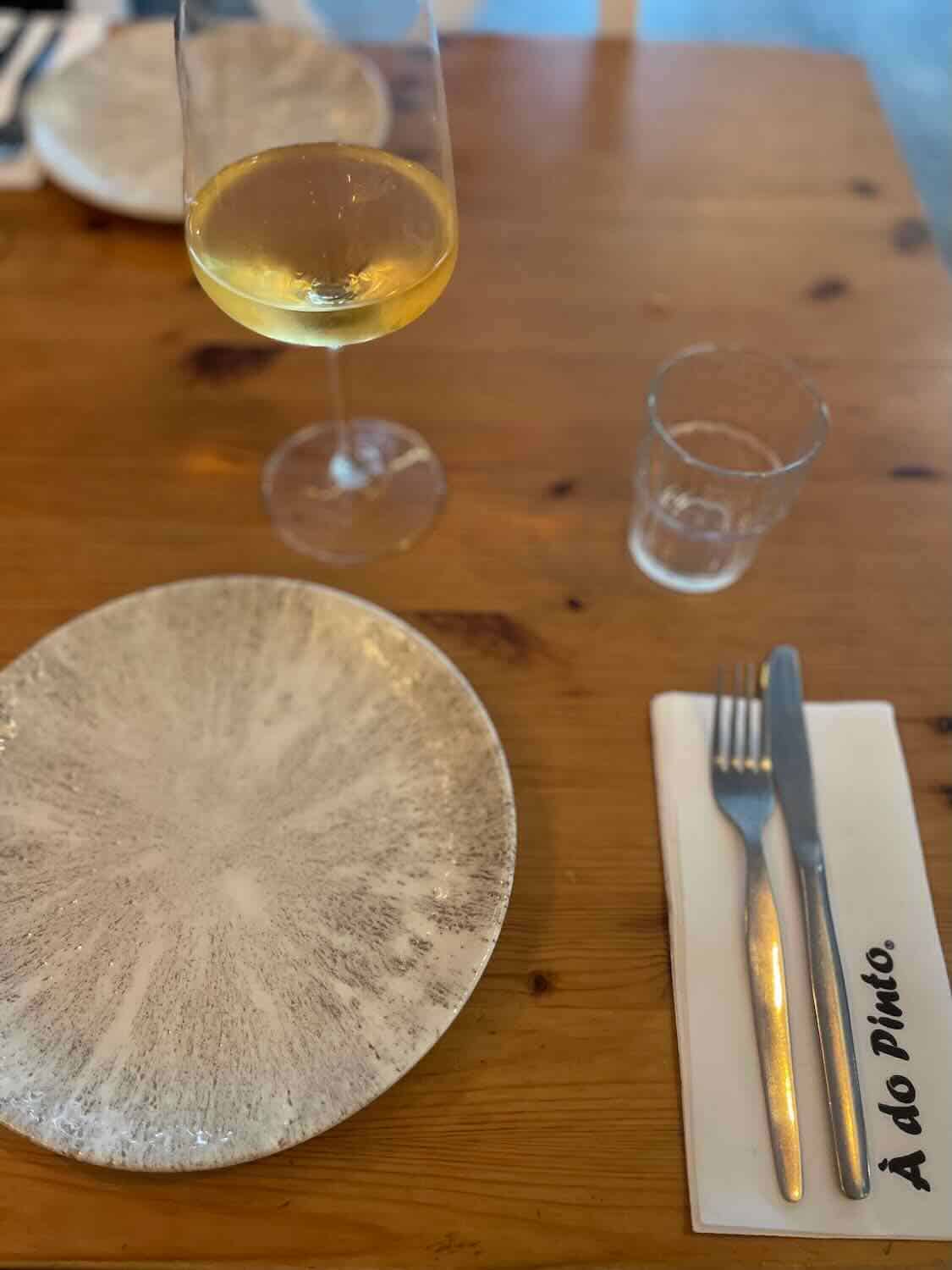 A glass of white wine on a wooden table beside a textured ceramic plate, with cutlery on a napkin reading 'A do Pinto,' suggesting an inviting dining experience.